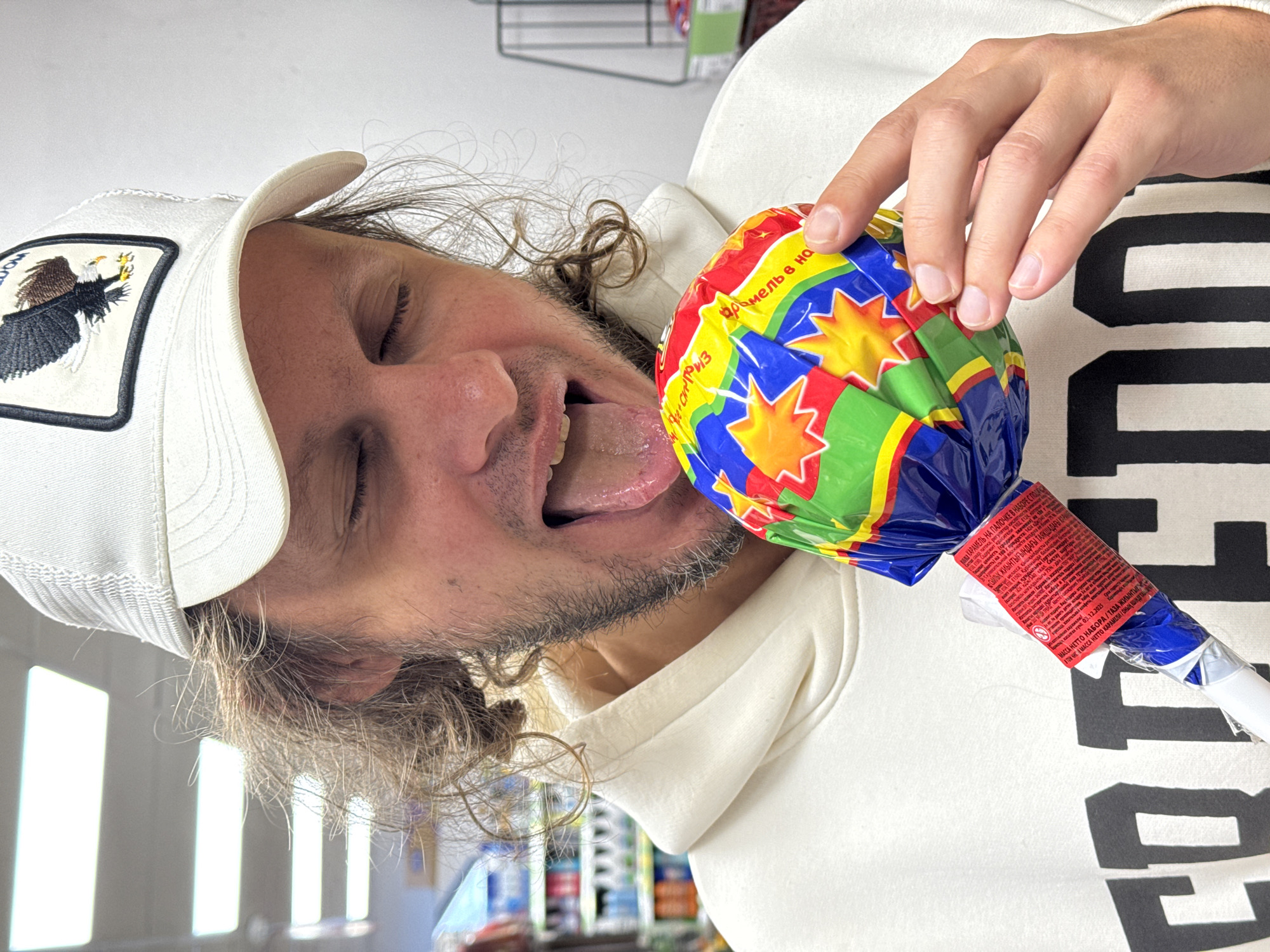 A fair-skinned man with shoulder-length wavy brown hair and a beard is holding a large, wrapped lollipop to his open mouth, with his tongue extended towards it. He is wearing a white baseball cap with an eagle patch and a white hoodie with a black graphic. The lollipop has colorful wrapping. The background suggests a retail environment, with shelves of products visible. The text on the lollipop is in Russian, indicating a location possibly in or near Novo-Nikol'skoe, Russia.