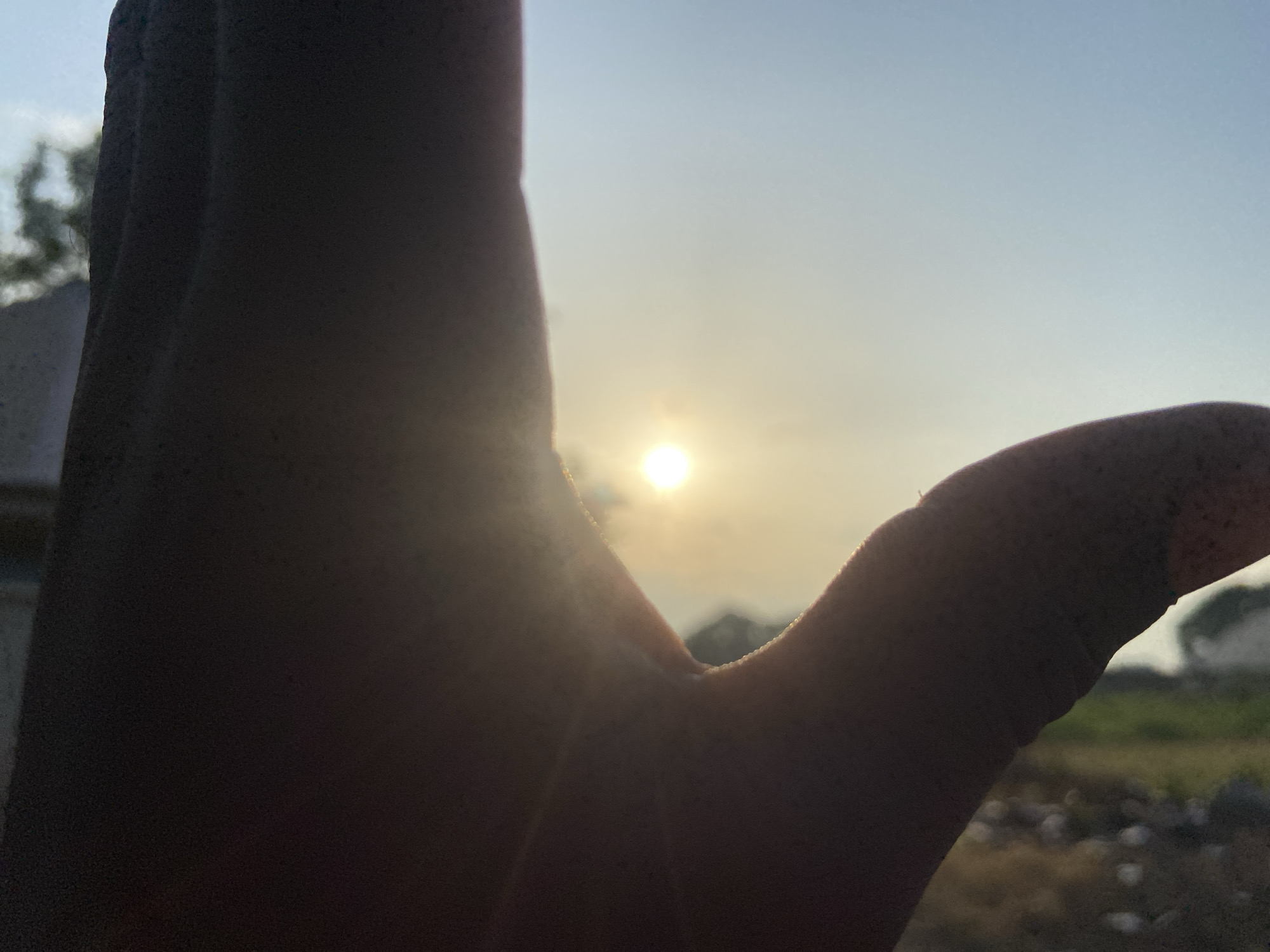 The media file captures a close-up view of a person's hand silhouetted against a bright sky at sunset. The sun is a small, glowing orb in the middle of the frame, partially obscured by the hand. The hand is positioned as if to cup or frame the sun. The background shows a hazy landscape with some distant trees and possibly buildings, suggesting an outdoor setting. The sky is a gradient of pale blue to a hazy yellow near the horizon, indicative of late afternoon or early evening. The overall impression is peaceful and contemplative, with the warm light of the setting sun creating a gentle atmosphere. The location is identified as Mararaba, Nigeria. There is no visible text in the media file.