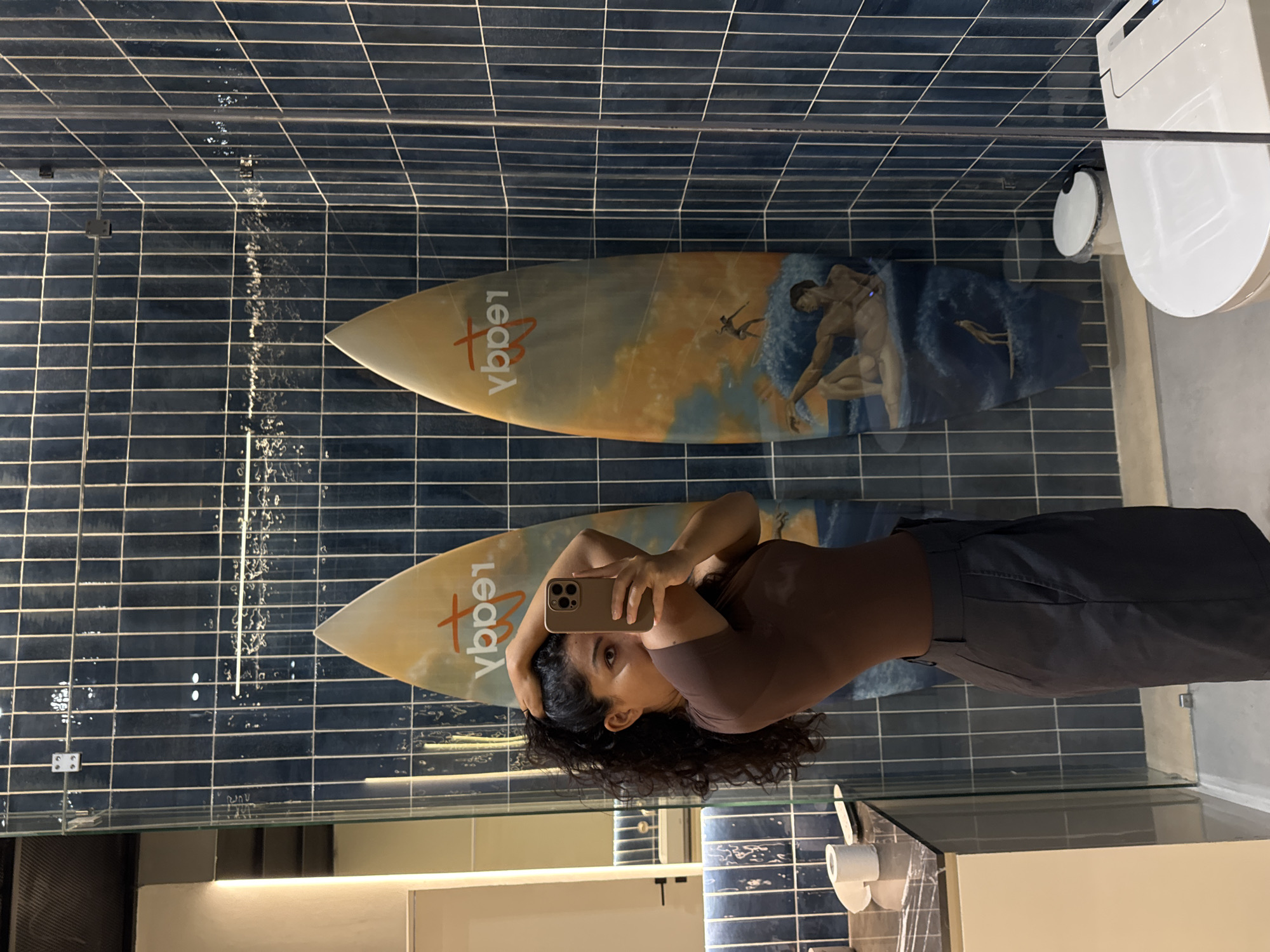 A woman with dark, curly hair, wearing a dark brown short-sleeved top and dark trousers, is captured taking a mirror selfie. She holds a light-colored smartphone to her face, obscuring part of it, while looking directly towards the camera with one eye visible.
The setting is a modern bathroom with dark blue rectangular wall tiles. Reflected in the mirror behind the woman are two surfboards leaning against the tiled wall. The prominent surfboard is light-toned, possibly beige or pale yellow, and features the word "Legacy" (visible as "ybpel" in the reflection) in a red, stylized script. It also displays a painted scene with muscular figures on ocean waves, reminiscent of classical art. A second, similar surfboard is partially visible behind it.
To the right side of the frame, a modern white toilet with a bidet function is visible. Below the mirror, a countertop or vanity holds a roll of toilet paper. A clear glass partition is also visible, indicating a separation within the bathroom space. The presence of surfboards, combined with the stylish and contemporary bathroom design, suggests a modern hotel or guesthouse setting, consistent with a location like Kuta Selatan, Indonesia. The indoor lighting does not indicate a specific time of day.
