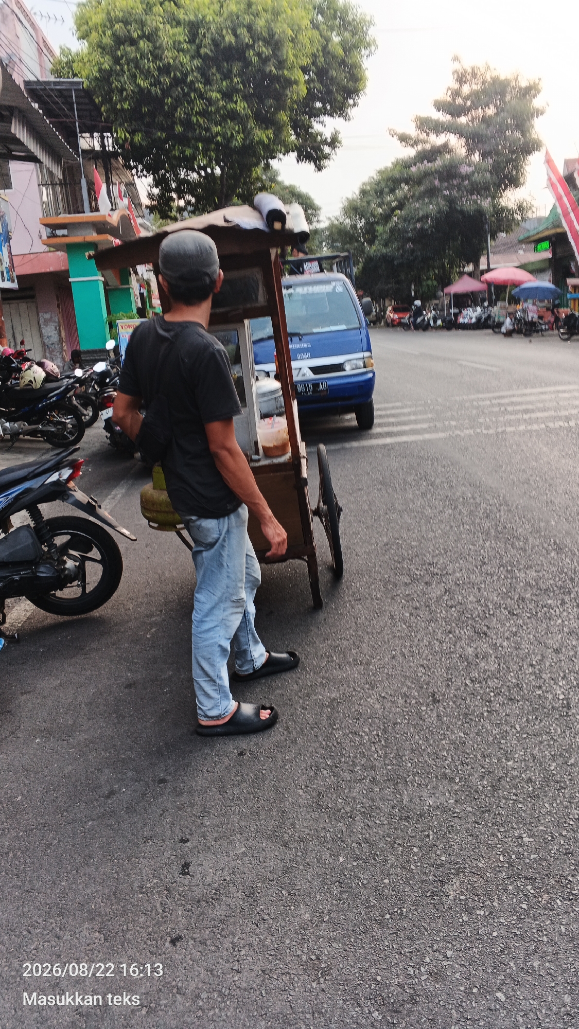 A person is pushing a food cart along a street in Malang, Indonesia. The person is wearing a dark t-shirt, light blue jeans, and black sandals. They have a gray cap on and a black bag slung over their shoulder. The food cart is made of wood with a metal frame and has a gas cylinder attached to the side. A blue van is parked nearby. In the background, there are buildings, trees, motorcycles, and other food carts with umbrellas. The scene appears to be taking place during the late afternoon or early evening. The timestamp on the image indicates the date August 22, 2026, at 16:13.