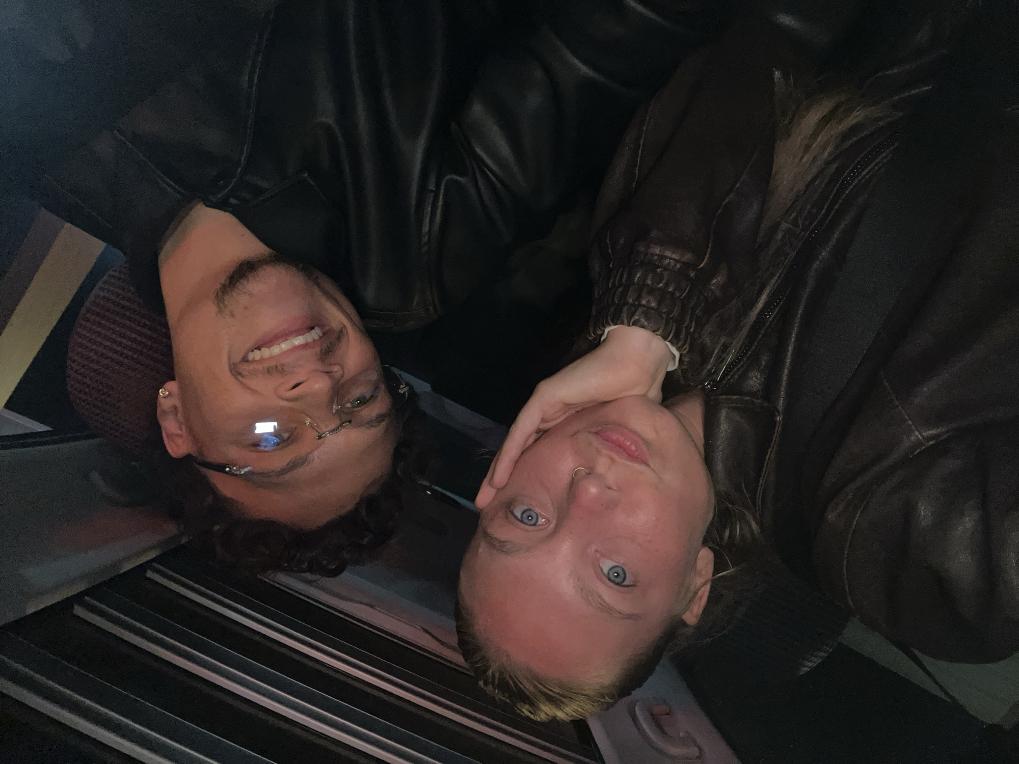Two individuals are visible in the image. On the left is a person with curly dark hair, wearing glasses and a black leather jacket. They are smiling, revealing their teeth. On the right, another person with light blonde hair and blue eyes is present. They are also wearing a leather jacket, but it is brown. Their hand is positioned near their face. The setting appears to be indoors, with a portion of a textured surface (possibly a seat) visible in the background. The presence of parallel lines suggests the inside of a bus or train. The location is unknown, but it has been requested that the city of Noisy-le-Grand, France, be mentioned. The image does not reveal the specific location of this scene within Noisy-le-Grand, France.