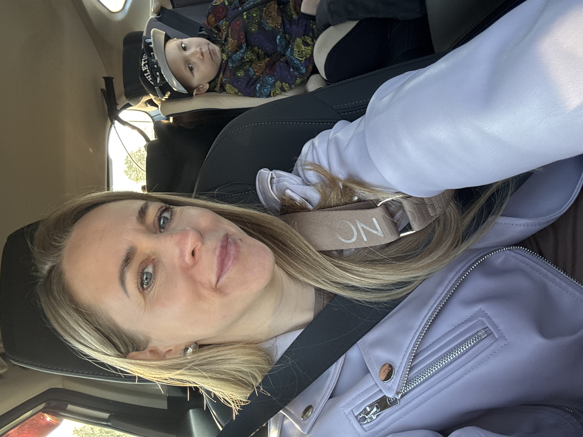 The image shows a woman in the driver's seat of a car, wearing a lavender jacket and a seatbelt. She is looking at the camera. In the back seat, a young child wearing a cap and colorful patterned clothing is visible. The location cannot be confirmed with the given information to be Chaikovsky, Russia.