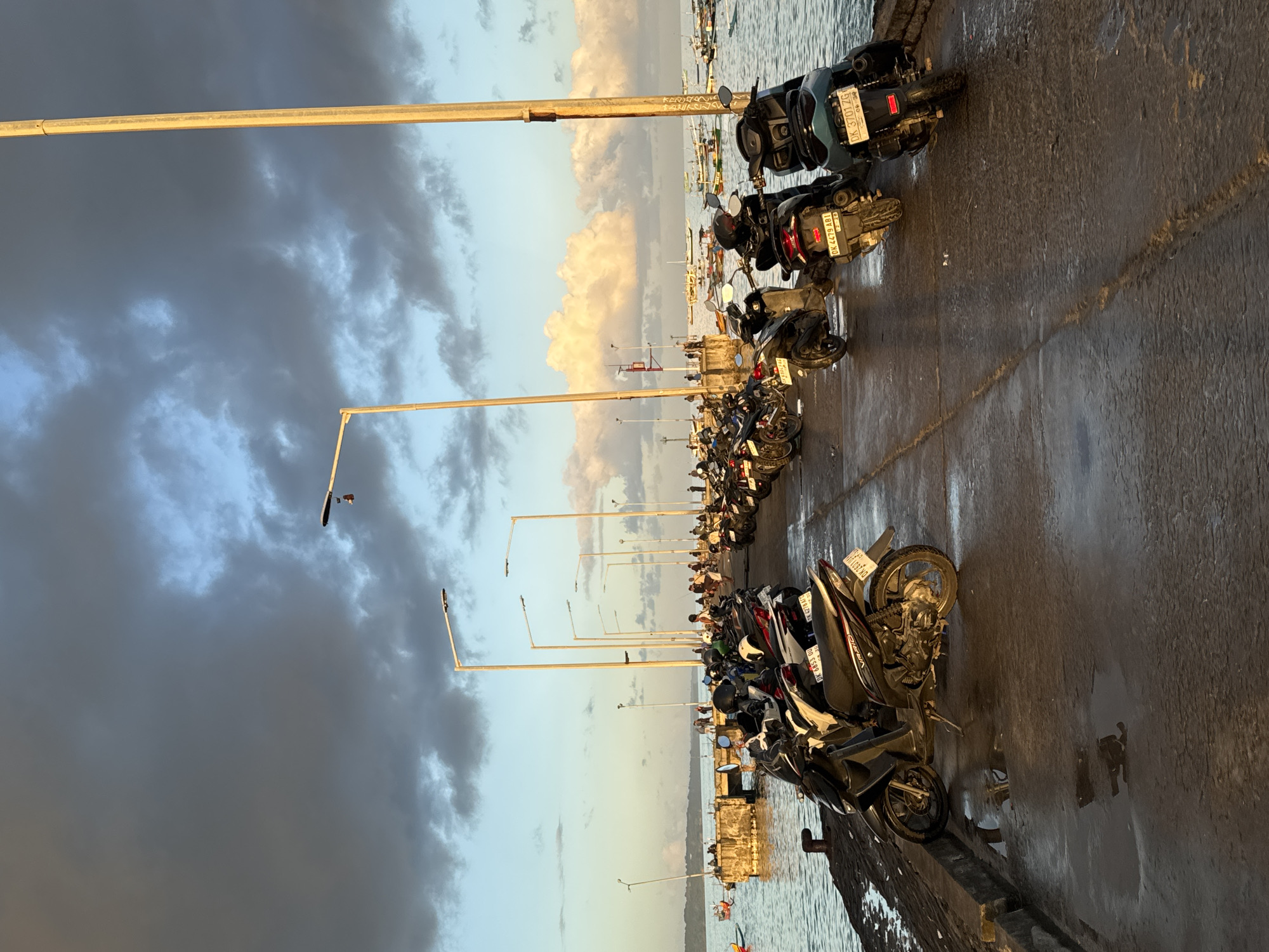 A long row of motorcycles is parked on a pier alongside the ocean. The pier is made of concrete and appears wet. Overhead, there are a series of tall, metal light poles extending down the pier. The sky above is mostly cloudy, with light blue visible near the horizon, indicating sunset or sunrise. In the distance, one can make out several boats on the water. The location appears to be a coastal area near Kuta, Indonesia.