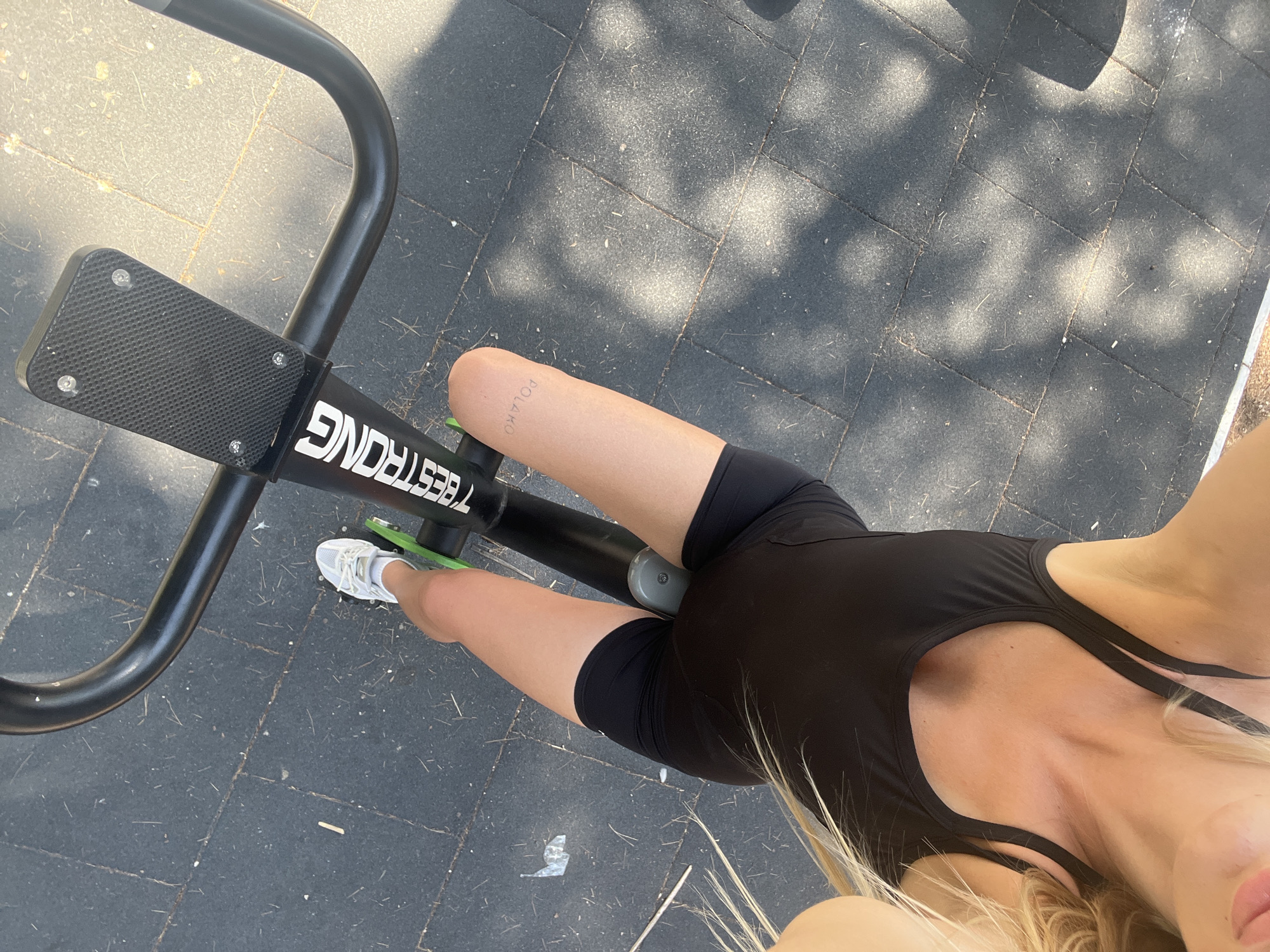 A high-angle, slightly overhead shot shows a person from the knees up, engaged in an outdoor workout in Bar, Montenegro. The subject is wearing black athletic shorts and a black one-piece athletic outfit. White athletic shoes are visible on their feet. The person is positioned on what appears to be an exercise machine, with a black frame and a textured black footplate that has "BESTRONG" written in white text. The ground is paved with dark gray, square tiles, and dappled sunlight creates a pattern of light and shadow on the surface, suggesting a sunny day. The background shows more of the tiled ground and hints of greenery or trees, indicating an outdoor setting. A faint tattoo is visible on the subject's thigh, with the word "POLAKO" written vertically. The overall mood is one of active fitness in a bright, outdoor environment.