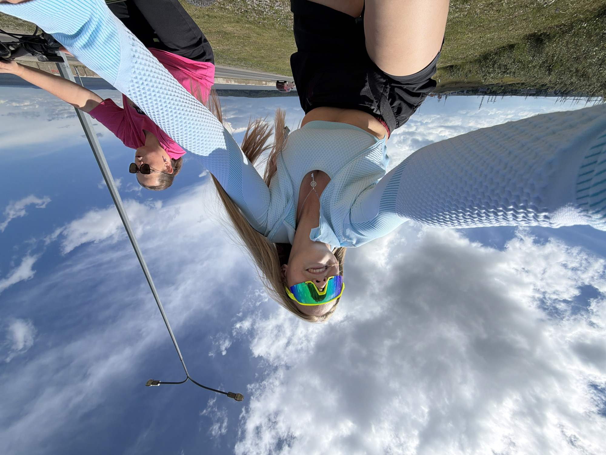 The image shows two young women in athletic wear, likely participating in an outdoor activity. One woman is wearing a bright pink top and black shorts, while the other is wearing a light blue long-sleeved top and black shorts. Both are wearing sunglasses. The woman in the light blue top is holding the camera and smiling, suggesting a lighthearted and fun atmosphere.
They appear to be outdoors, with a grassy area and what looks like a road or track in the background. The sky is visible, filled with clouds, and a street lamp is also present. The overall impression is that of a recreational or sporting event taking place during the daytime, possibly on a clear day given the brightness of the scene. The image is taken from a low angle, making the sky a prominent feature. The women's clothing suggests they are active and dressed for warm weather. There is no visible text in the image.