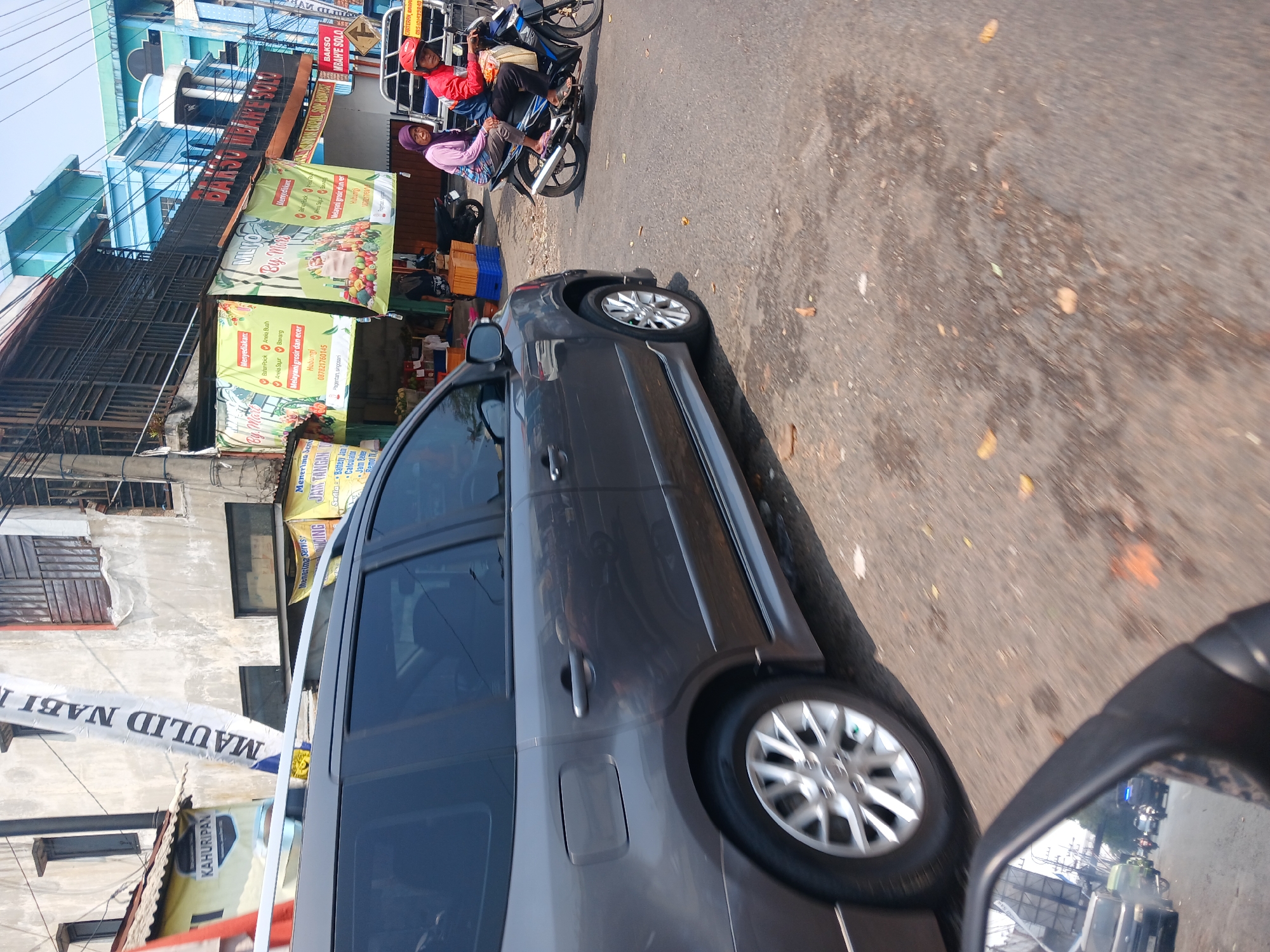 The image depicts a street scene in Singosari, Indonesia, on a sunny day. In the foreground, a dark gray minivan is parked or moving slowly on the asphalt road. The minivan has silver multi-spoke wheels and a roof rack. A reflection of traffic, including a black van and motorcycles, is visible in the side mirror of a vehicle in the foreground, which is partially captured at the bottom left of the image.

Behind the minivan, there are several buildings and businesses. One building has a weathered gray facade with visible windows and some wooden-slatted details. A white banner with blue text, reading "MAULID NABI," is prominently displayed vertically on this building. Adjacent to it, a yellow banner with the text "KAHURIPAN" is visible.

Further down the street, a row of shops features colorful banners. One banner, predominantly green, advertises "MLRO BY. MAS" and lists items like "Menyediakan: Bawang Putih," "Aneka Buah," and "Melayani grosir dan ecer." Another banner, also green, appears to be similar. A red sign with white text, "BAKSO MBAH E SOLO," is mounted on one of the buildings.

On the road, a man wearing a red jacket and a red helmet is riding a motorcycle, with a woman wearing a purple headscarf seated behind him. They are carrying a white bag. Other motorcycles and a silver three-wheeled vehicle are also present on the road. The street is paved with asphalt and shows signs of wear and some patches of wetness, with scattered leaves. The overall impression is of a bustling urban environment.