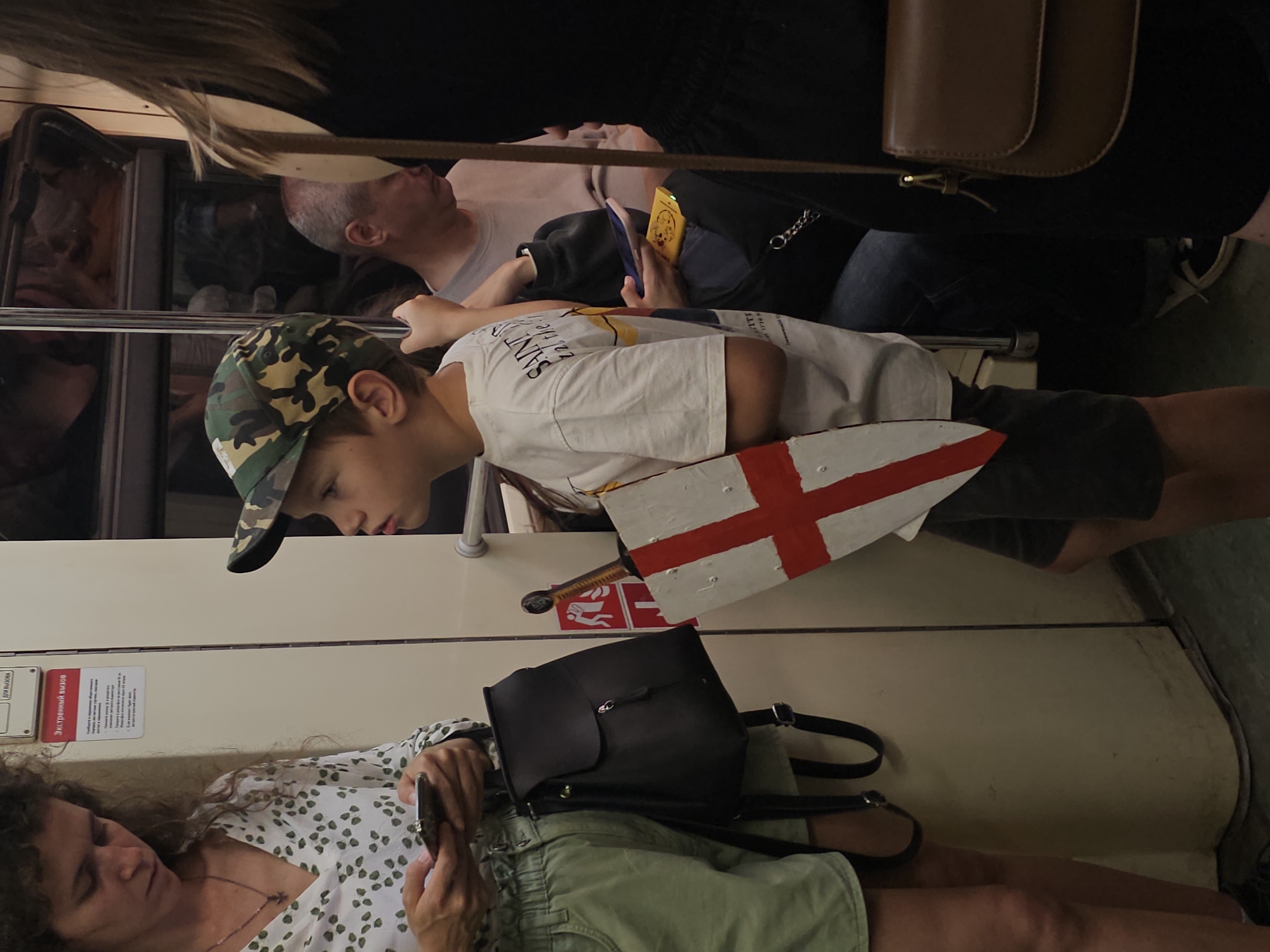 A boy wearing a camouflage baseball cap and a t-shirt stands on a subway train, holding a toy sword and shield painted with the English flag. To his left, a woman with curly hair, wearing a green patterned blouse and light-colored shorts, looks at her phone. Behind the boy, other passengers are visible, including a man with gray hair wearing a t-shirt and a person in a black top holding a phone. The environment appears to be the interior of a subway car with cream-colored walls and metal poles.