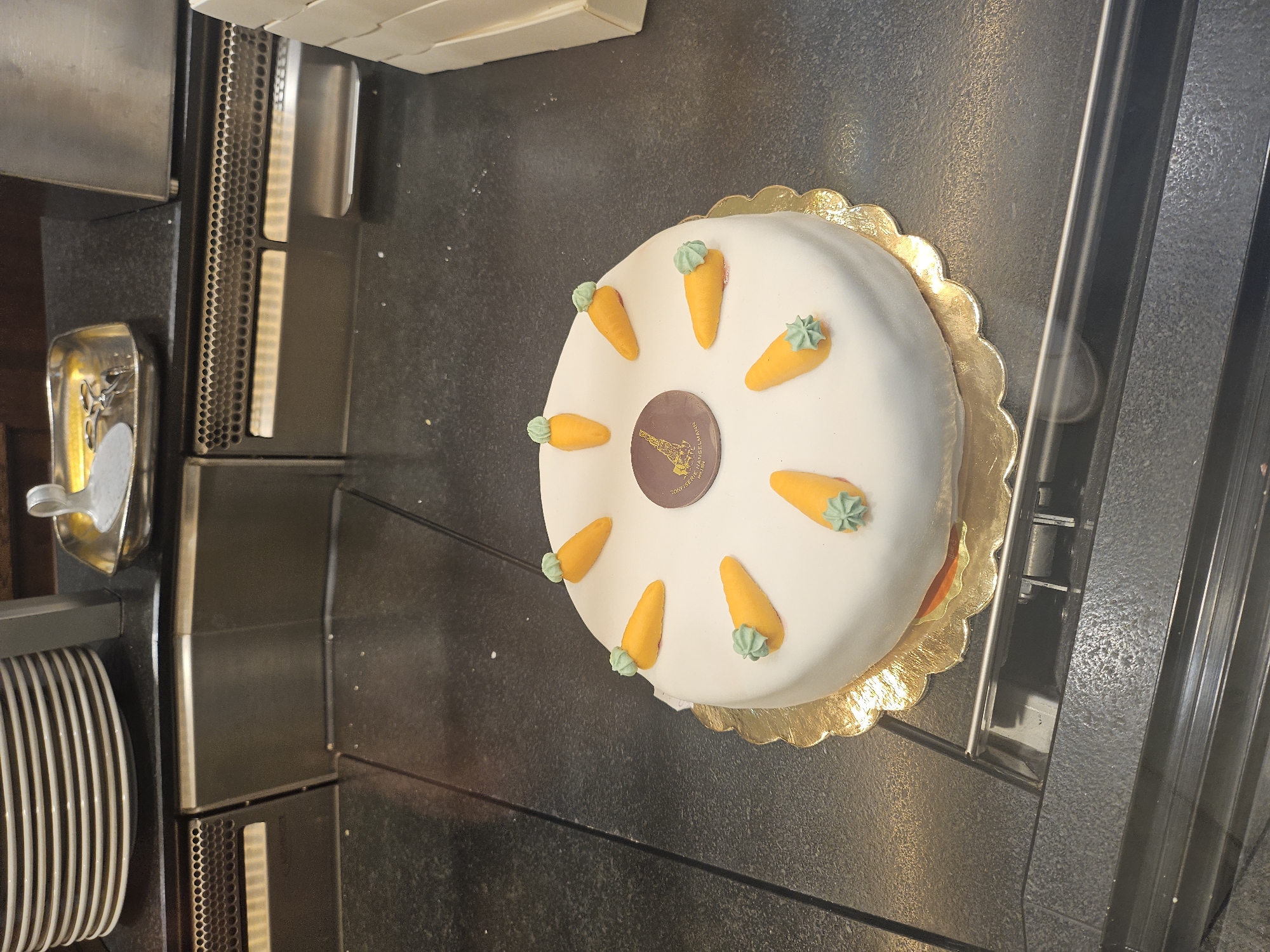 A white frosted cake, decorated with nine small orange carrot-shaped candies with green frosting tops, sits on a gold scalloped cake board on a black countertop. The cake is round and has a dark brown circular emblem in the center, depicting a crane and text that appears to read "Konditorei Sprüngli Zürich". The countertop is speckled and appears to be in a commercial setting, possibly a bakery or cafe, with stacked plates visible in the background to the left and a stainless steel apparatus to the right. The overall impression is of a well-presented dessert item. The location of this scene is the Konditorei Sprüngli in St. Moritz, Switzerland, a renowned confectionery shop established in 1859.