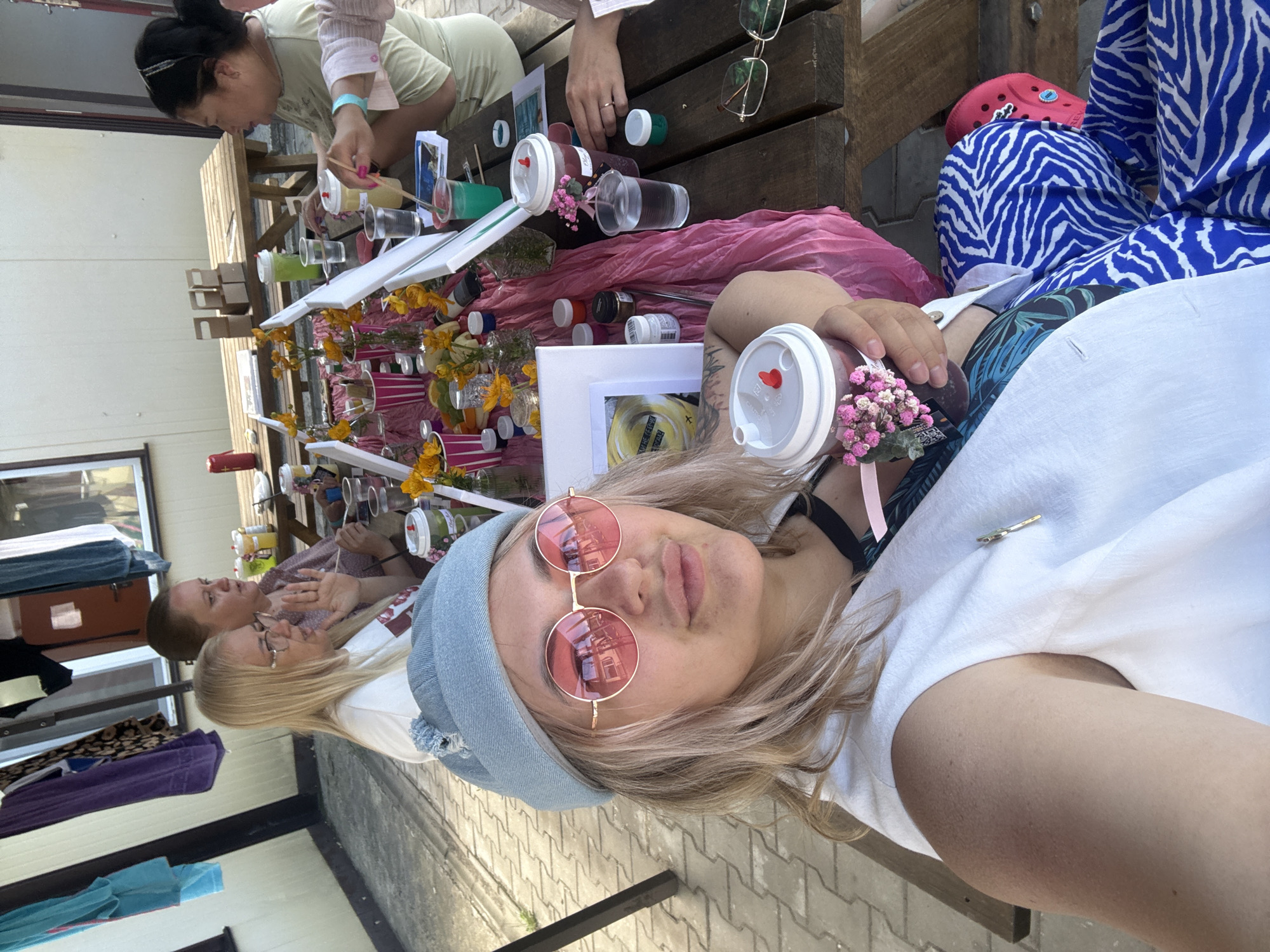 In Birobidzhan, Russia, a group of individuals are gathered around a wooden picnic table outdoors, possibly for a creative activity. The table is adorned with a pink fabric runner, small vases with yellow flowers, fruit, and various art supplies including paint pots and brushes. Two women are visible at the table, one with blonde hair wearing glasses and a white shirt, and another with dark hair wearing a light green shirt, holding a brush and looking towards the table. In the foreground, a woman with blonde hair, wearing a light blue beanie and round pink-lensed sunglasses, takes a selfie while holding a drink adorned with small pink flowers. She is wearing a white vest over a patterned top and blue and white zebra-print pants. The background shows a building with clothes hanging on a line, and a paved walkway.