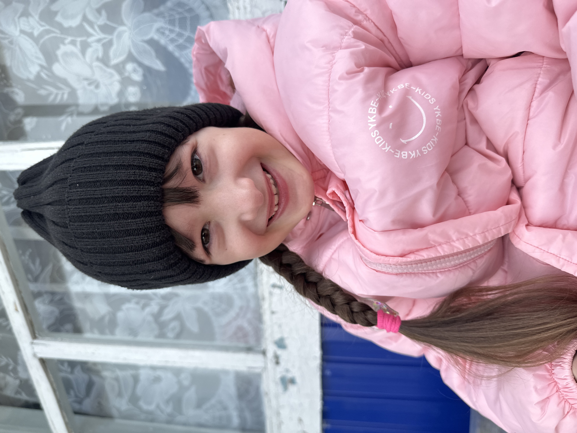 A frontal portrait depicts a young female child in Aktanysh, Russia, positioned against a background of a window and an adjacent wall. The child has fair skin, dark brown hair styled with bangs and a long braid secured by a pink elastic band and a translucent accessory on her left side. She wears a black ribbed knit beanie and a pink puffer jacket. The jacket displays a white circular logo on the left chest area, featuring a smiley face surrounded by "YKBE-KIDS" text. The child exhibits a wide smile, revealing teeth, with her eyes open and directed towards the camera. The immediate background behind the child consists of a weathered white wooden window frame with multiple panes. Through these panes, a translucent white material with a floral pattern is visible, consistent with a curtain or frosted glass. Below the window, to the lower left, a segment of a blue corrugated or paneled surface is present.