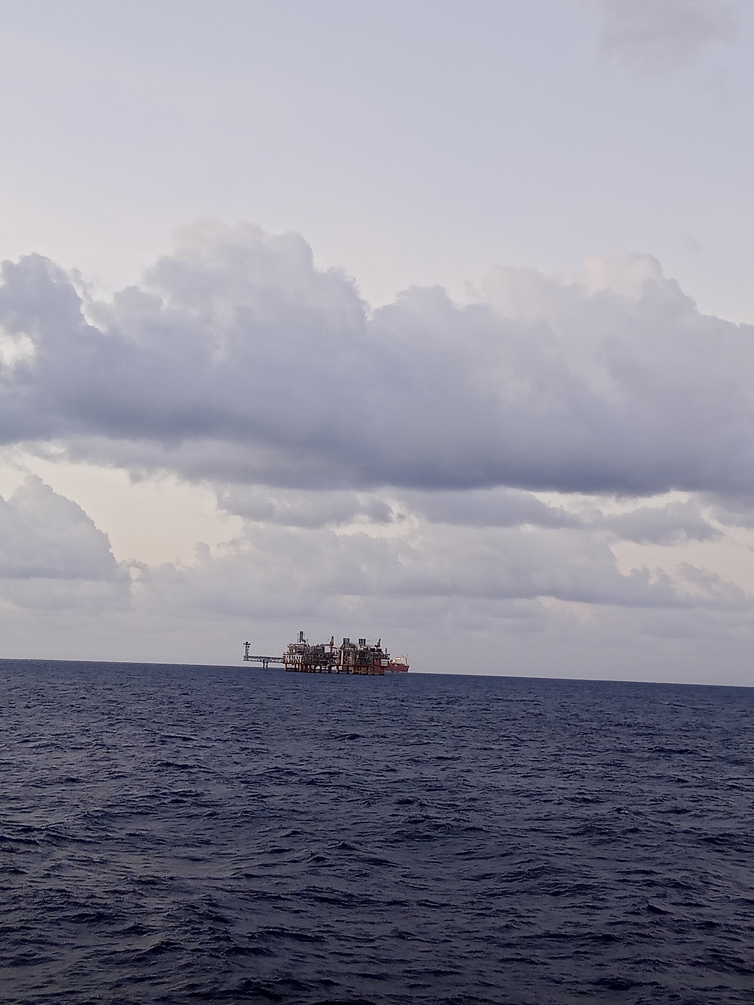 An oil rig is situated in the middle of the ocean under a cloudy sky. The water is dark blue and appears choppy. The oil rig is a complex structure with multiple levels and a connecting bridge.