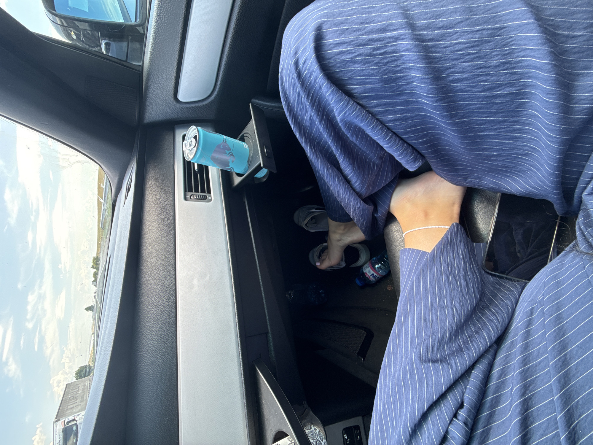 The image shows the interior of a car on a highway, with a view of the road and sky through the windshield. The focus is on the passenger's legs and feet, wearing blue and white striped pants. A can of Red Bull is in a cup holder, and two water bottles are on the floor. The overall setting suggests a journey on a sunny day. The location is identified as Polsko Padarevo, Bulgaria.