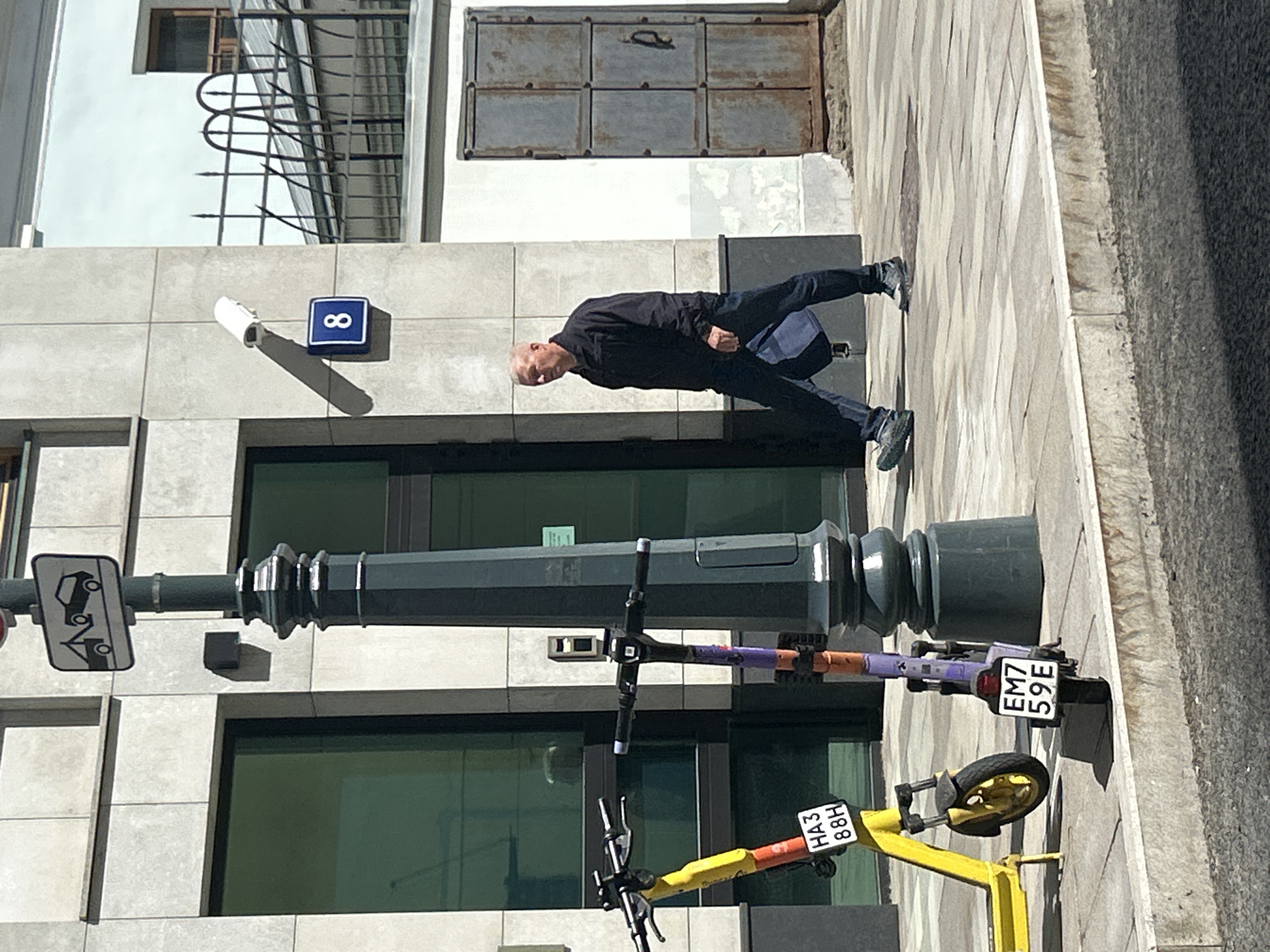 A man in a black jacket and dark pants walks on a sidewalk in front of a building with large windows. He is carrying a blue bag. Two electric scooters are parked on the sidewalk to the left of the man. One scooter is yellow and the other is purple. A street light with a tow-away sign is visible on the left side of the image. A blue sign with the number "8" is mounted on the building. A metal door is visible on the right side of the image. The sidewalk is made of concrete pavers, and the street is paved with asphalt. The scene appears to be in an urban environment.