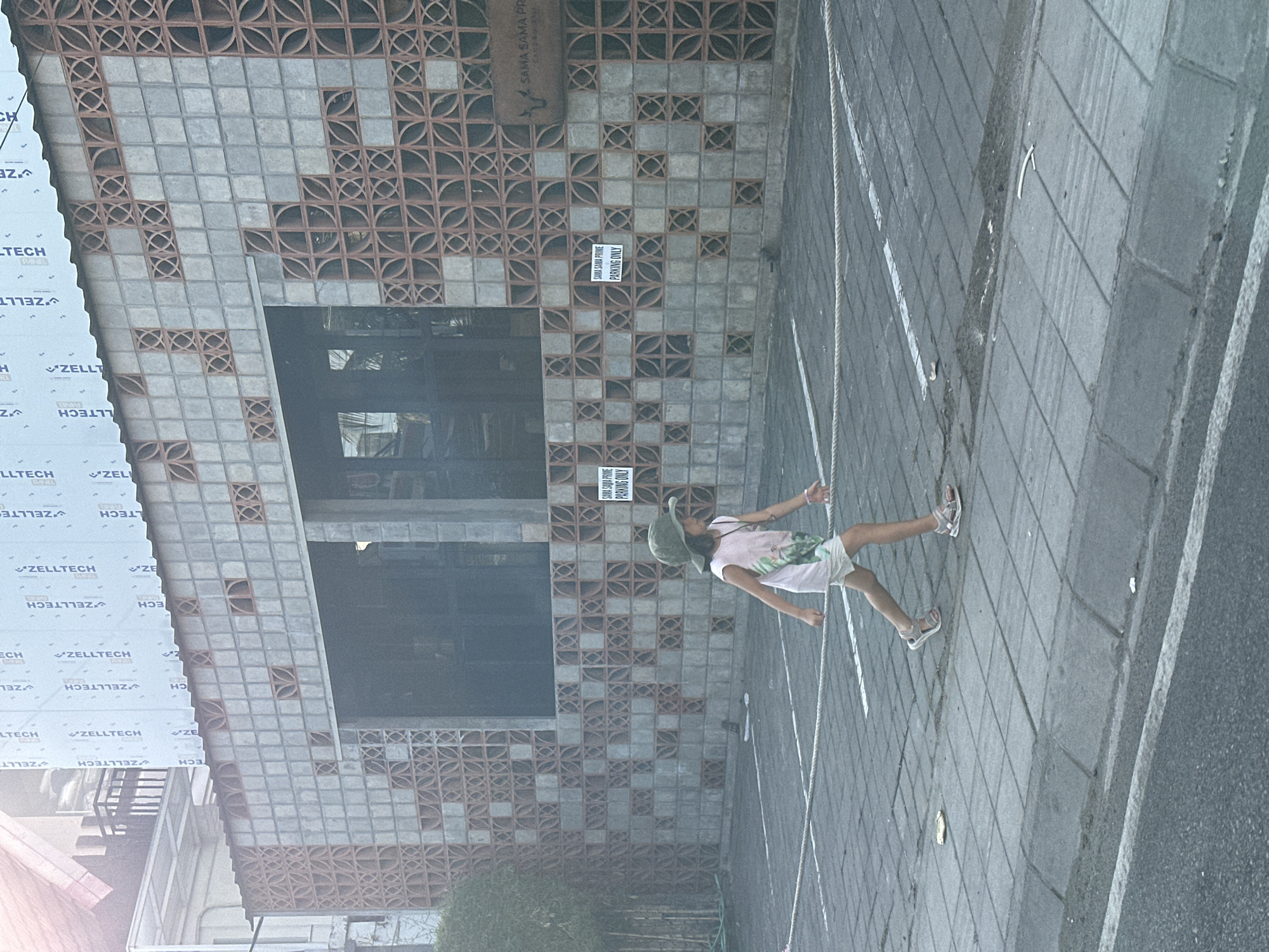 A young girl wearing a light-colored t-shirt, white shorts, a green bucket hat, and sandals walks on a paved walkway adjacent to a building. The building features a distinctive wall constructed with patterned concrete blocks, some of which are square and others circular. There are visible signs on the building, including one that reads "SAMA SAMA PRIME PARKING ONLY." The walkway is marked with white lines, and a rope barrier is present. The environment appears to be an outdoor urban setting.