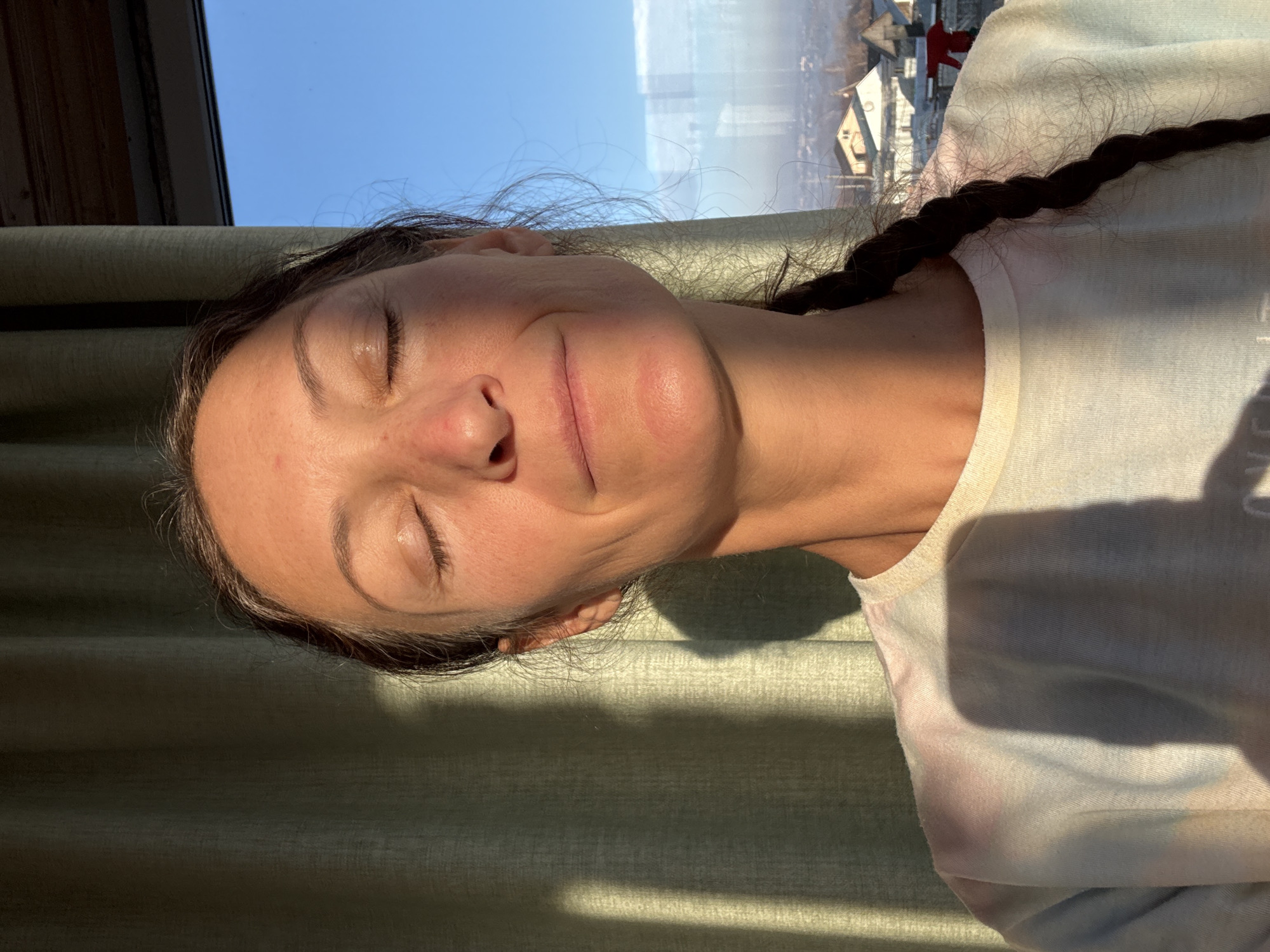 A woman with closed eyes and a braided hairstyle is positioned against a light green curtain. The lighting is bright, casting shadows on her face and the curtain. In the background, a window reveals a cityscape with houses, possibly located in Ulyanovsk, Russia. She is wearing a light-colored top.