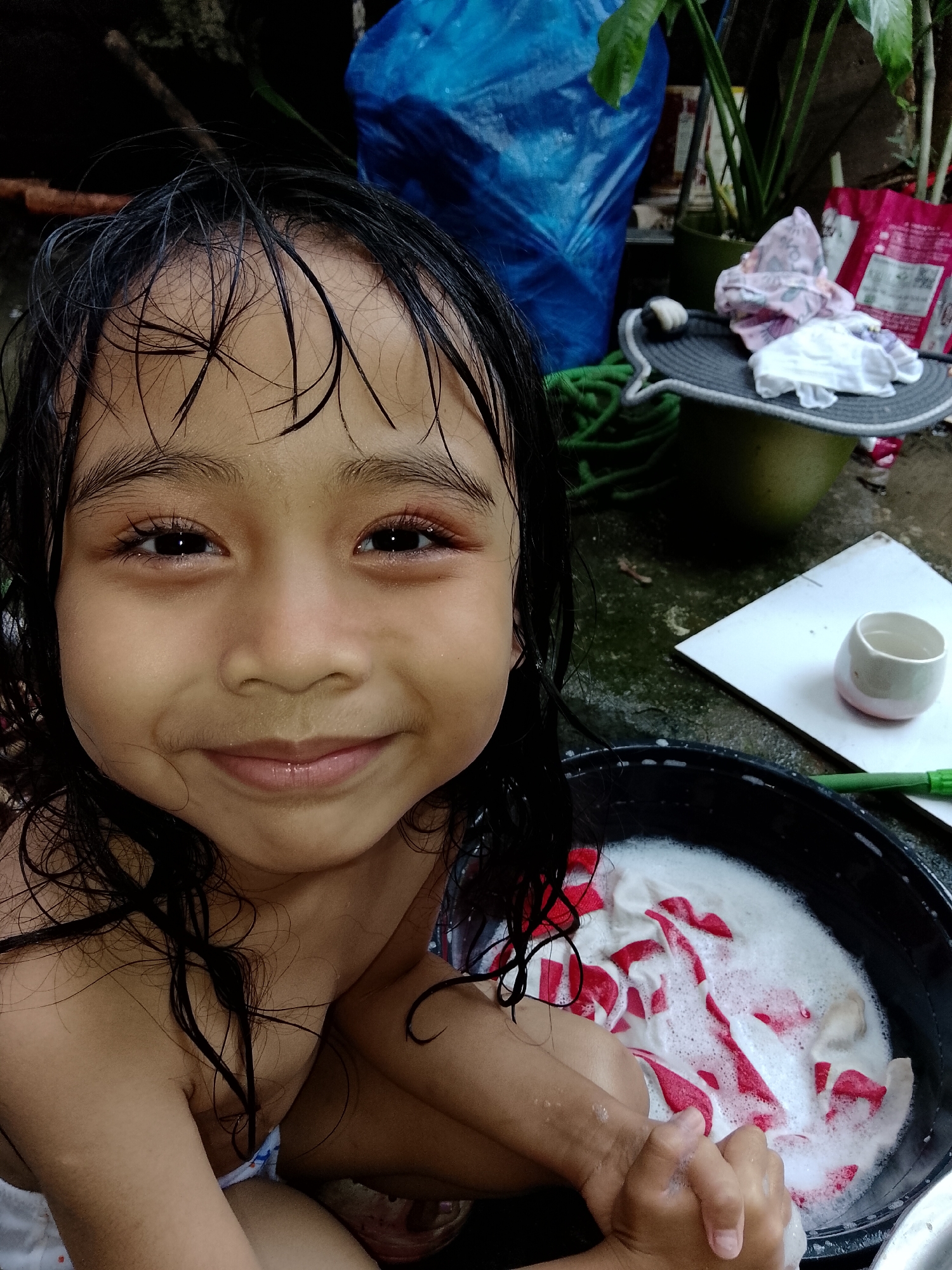 A young Asian girl with wet, dark hair and a warm, closed-mouth smile looks directly at the viewer. Her bare shoulders are visible, and she is crouched, hand-washing laundry in a black plastic basin filled with white soap suds and red-and-white striped fabric. Her hands are submerged in the soapy water. The setting is an outdoor, informal domestic area in Davao City, Philippines, characterized by a damp concrete or tiled ground. In the background, from left to right, are observed: dark soil or debris, a large blue plastic tarp, a coiled green hose, and a potted plant with broad leaves. To the right of the basin, a small white ceramic cup with a pinkish base rests on a white tile, next to a green-handled brush or tool. Further right, a grey woven hat with a white band is perched on a green bucket, adorned with folded light-colored garments. A partially visible pink package or bag is in the far right. Natural daylight illuminates the scene, highlighting the wet surfaces and the child's clear, dark eyes.
