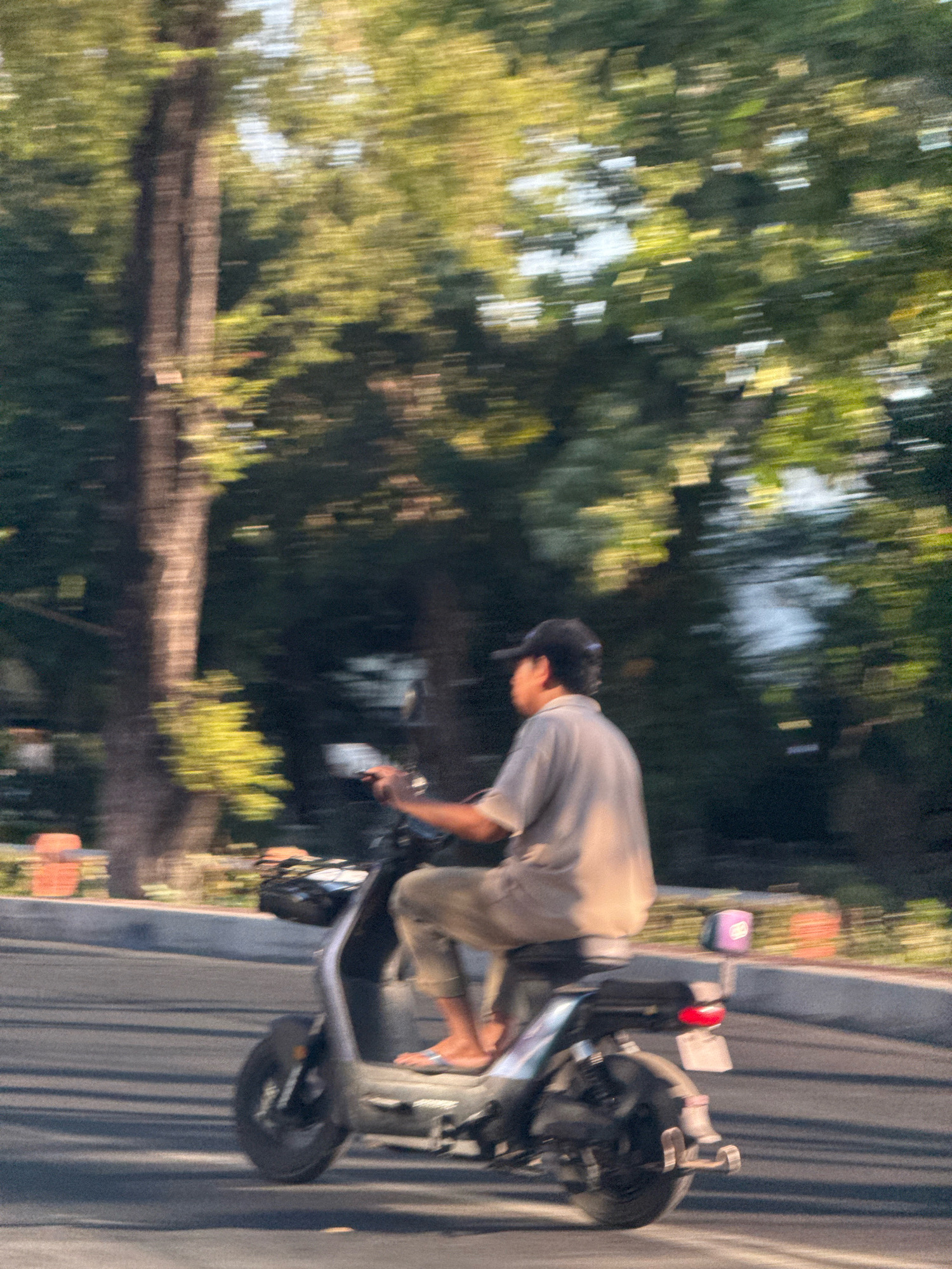 A person wearing a cap, grey shirt, and light-colored pants rides an electric scooter. The scooter is silver and grey, with a basket at the front and a red taillight. The person is seen from the side, with their feet on the footrest. The scooter is on a paved road with some debris and shadows. In the background, there are trees with green and yellow foliage, suggesting a sunny day. The motion blur indicates the scooter is moving. The scene is set in a city environment, with the presence of a road and trees lining it. Based on typical scenery in Surabaya, Indonesia, this could be a street with mature trees and urban infrastructure.