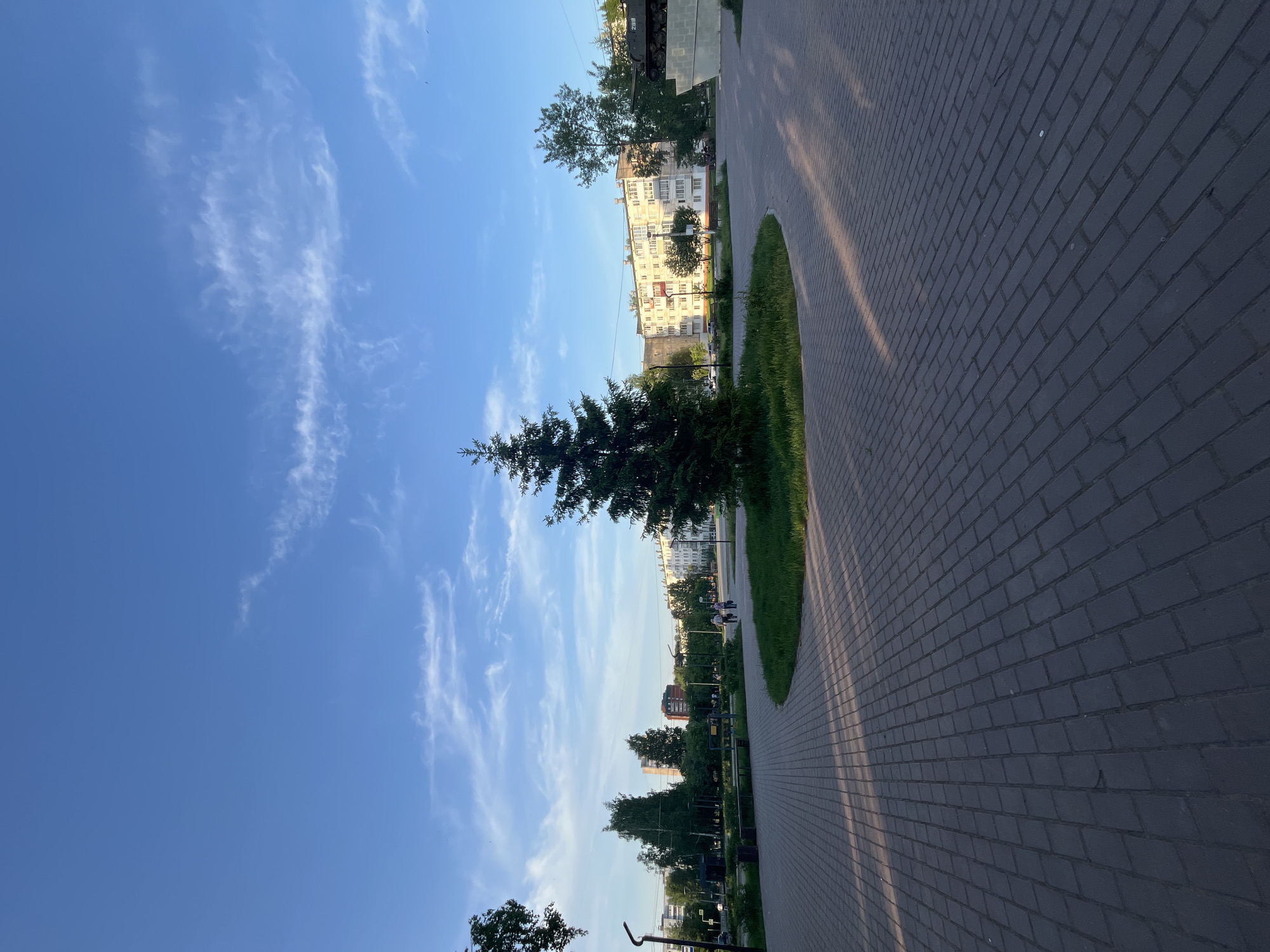 The image captures an urban scene in what appears to be a residential area. A paved brick walkway, laid out in a herringbone pattern, occupies a large portion of the foreground. The path is bordered by a strip of grass and low vegetation. A tall evergreen tree stands prominently along the walkway.
In the background are multi-story residential buildings of Soviet-era architecture, painted in light colors. The sky above is blue with wispy clouds. The image is slightly tilted, presenting the scene from a skewed perspective.
Based on the architectural style and general environment, the location is likely in Chaikovsky, Russia. The photo appears to be taken in the evening or early morning as long shadows are visible across the brick pathway.