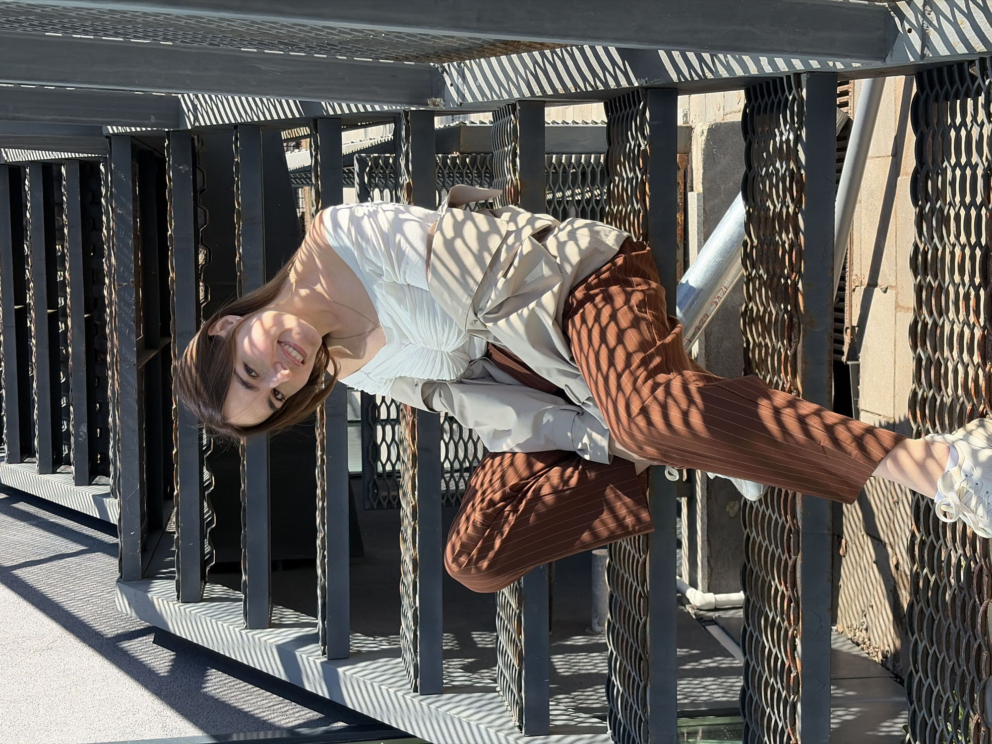 A young woman with long brown hair is posing upside down while sitting on a metal railing. She is wearing a white top, a beige jacket, and brown striped pants with white sneakers. The location appears to be an elevated walkway with metal grate flooring and railings, possibly a pedestrian bridge or fire escape. The harsh sunlight casts shadows through the railing structure. The location in the city of Izhevsk, Russia, is not visually discernible from the image alone.
