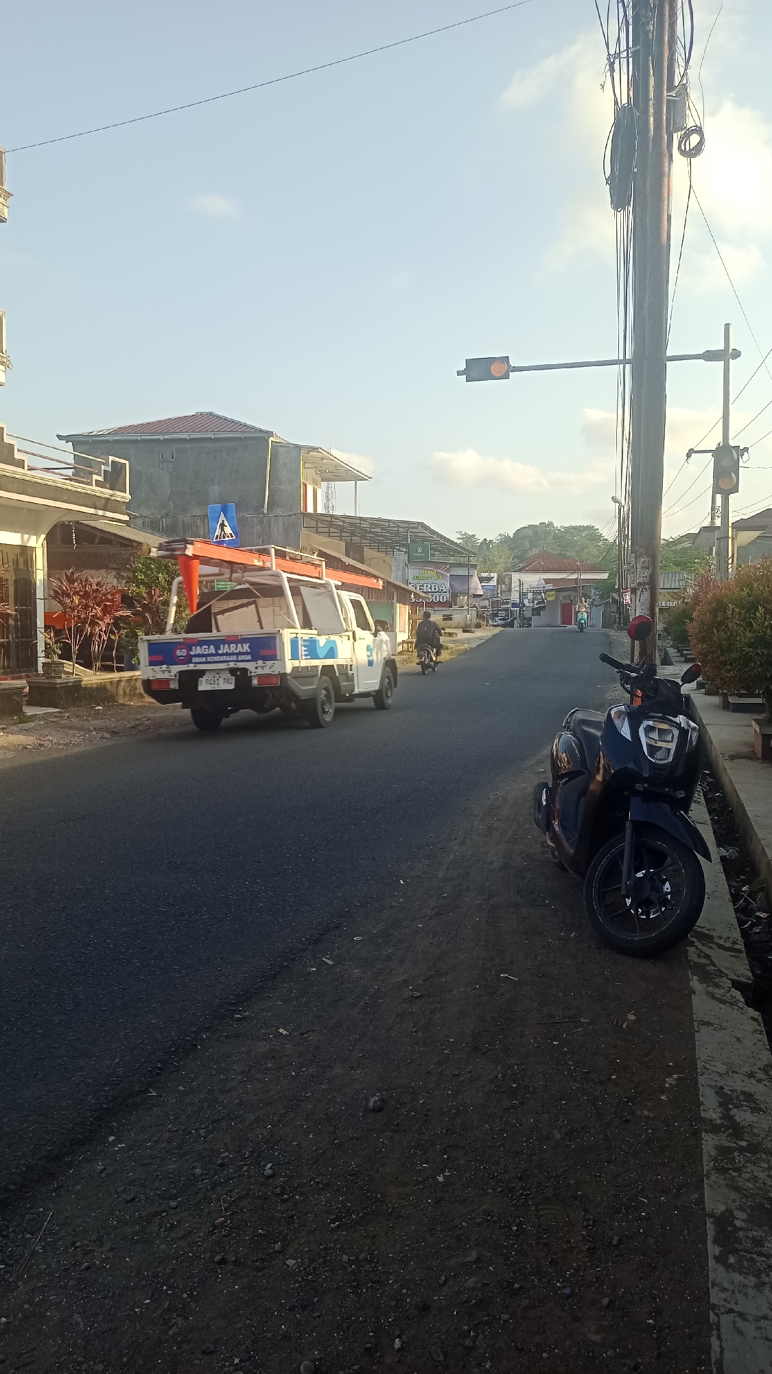 The image depicts a street scene in Karyamandala, Indonesia, under a clear sky with scattered clouds. In the foreground on the right, a dark-colored scooter is parked on the side of the road, facing left. The road itself is paved and appears to be a two-lane street.
On the left side of the road, a white pickup truck is parked, facing away from the camera. The truck has an orange ladder rack on its roof and displays markings on its side that include "60 JAGA JARAK" and "AMAN KENDARAAN ANDA". The truck appears to be loaded with equipment.
Further down the street, several buildings are visible, including what appear to be shops or houses. A traffic light is positioned on a utility pole on the right side of the road, and another traffic light is visible further in the distance. A few individuals are seen on motorcycles in the mid-ground. The environment appears to be a semi-urban or rural setting, with vegetation visible along the sides of the road and in the background. The lighting suggests it is daytime, possibly late afternoon or early morning.