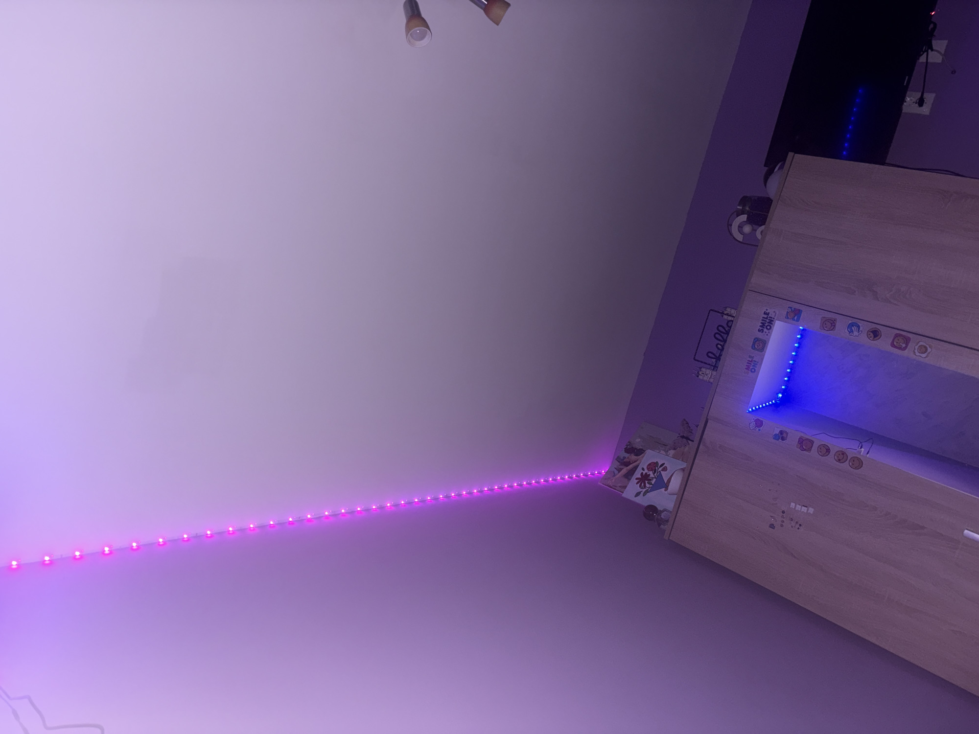 The image depicts an interior room with walls painted a light purple color, illuminated by a string of pink LED lights that run vertically along the corner of the room from the ceiling downwards. In the lower portion of the image, a light-colored wooden wardrobe is visible. The top of the wardrobe is adorned with various decorative items, including a framed picture with flowers, a small globe, and a miniature castle. The wardrobe doors have some decorative stickers, and one of the doors has an opening that reveals a blue LED light strip. A dark-colored television is mounted on the wall to the right of the wardrobe. The scene is likely a bedroom or a personal living space. The image does not contain any information that would allow for its location to be determined within Kirilovo, Bulgaria.