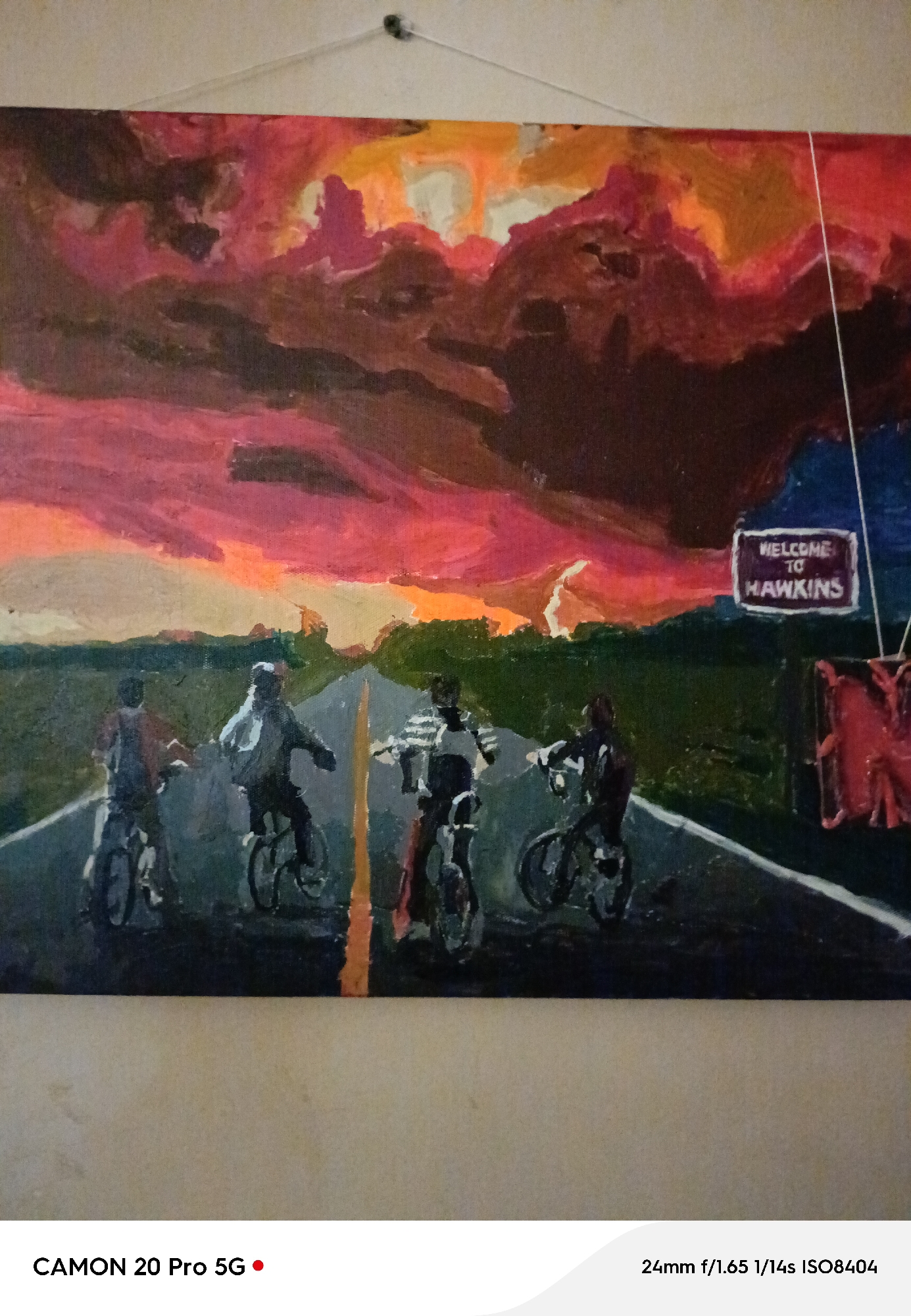 The image is a photograph of a painting that depicts four figures on bicycles riding on a road. The sky in the painting is a dramatic blend of red, orange, and dark hues, suggesting a sunset or stormy weather. A sign that reads "WELCOME TO HAWKINS" is visible on the right side of the painting. The painting is hanging on a textured wall. Below the painting, there is text indicating "CAMON 20 Pro 5G" and camera settings "24mm f/1.65 1/14s ISO8404". The painting does not depict any recognizable landmarks or features of Yakutsk, Russia, and therefore, its location within the city cannot be determined.
