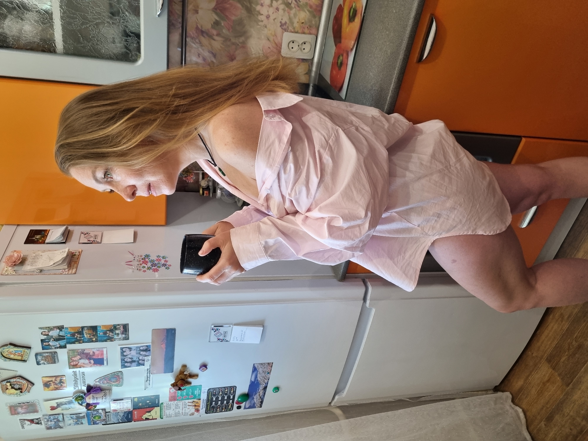 A woman with blonde hair is standing in a kitchen, wearing a pink button-down shirt and seemingly no pants. She is holding a black mug in her hands. Behind her, there is a refrigerator covered in magnets and photographs, with orange kitchen cabinets above it. The floor appears to be wood. Based on the available information, there is no indication that this image was taken in the city of Chaikovsky, Russia. More information would be needed to verify this aspect of the query.