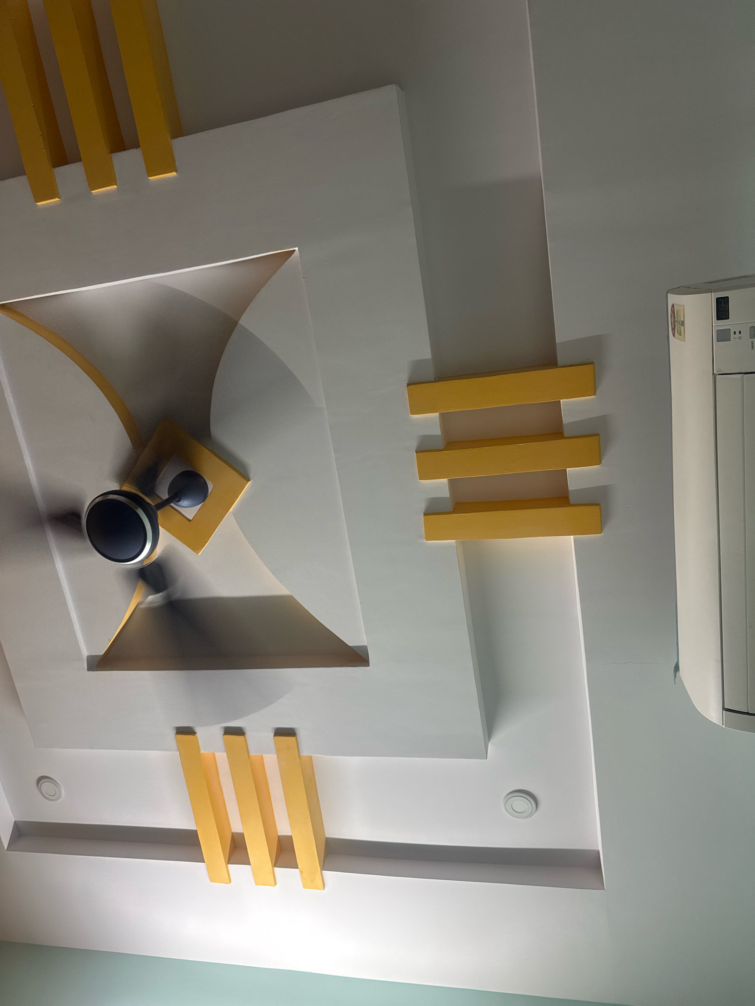 An interior shot taken from a low angle looking up at a modern ceiling design. The ceiling features multiple levels and geometric patterns in white, accented with bright yellow rectangular elements. A black and silver ceiling fan is centrally located within a recessed square area, suspended from the ceiling. To the right, a white air conditioner unit is mounted on the wall. The scene is well-lit, highlighting the architectural details of the ceiling. The image appears to be taken inside a residential or commercial space. Based on the metadata, the location is Nagamalli Thota, India.