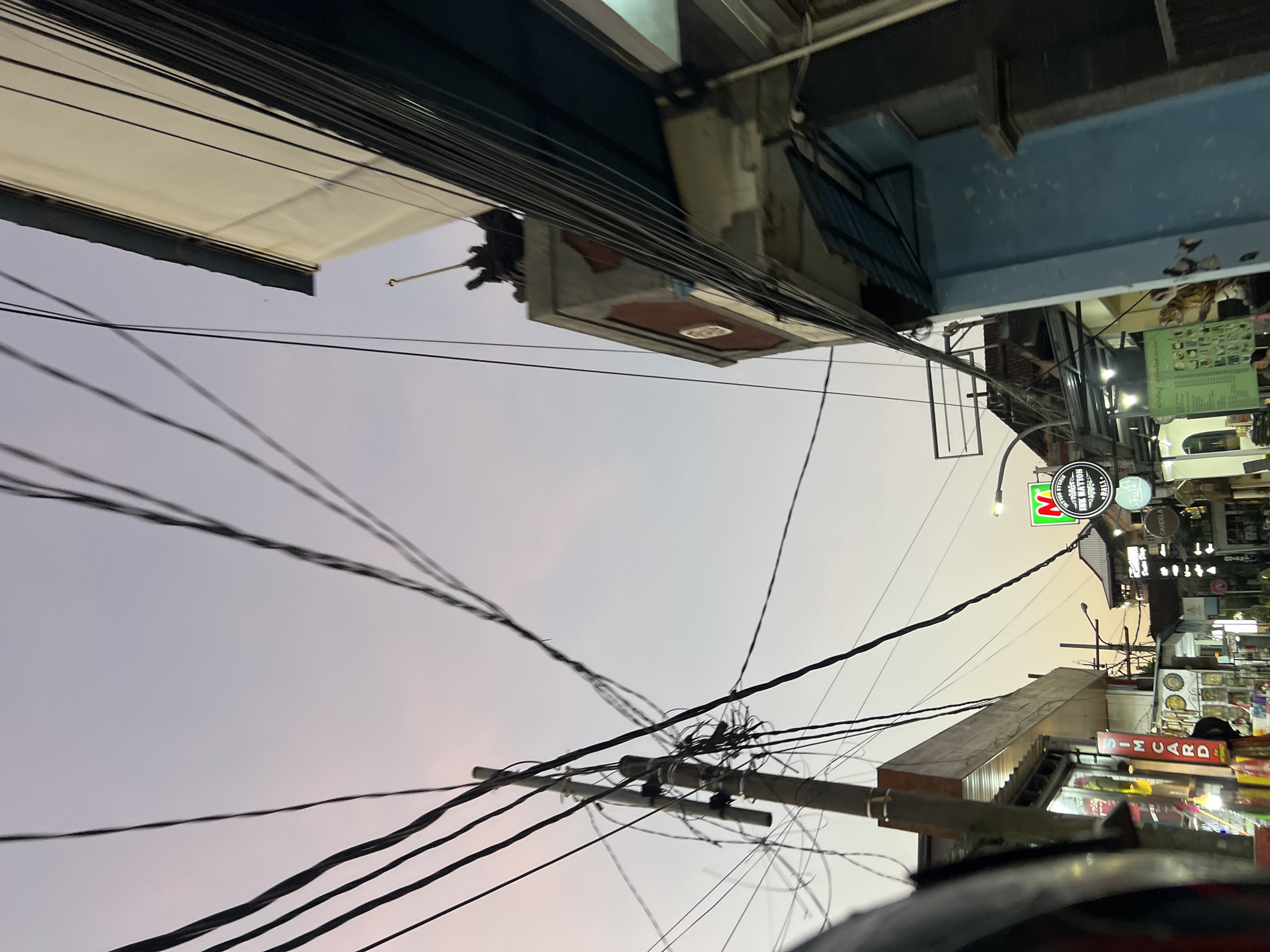 The image shows a street scene in Kuta, Indonesia, likely during dusk or dawn. The sky is a soft gradient of pink and pale blue, indicating the time of day. A tangled network of electrical wires dominates the upper portion of the frame, crisscrossing against the sky and obscuring the view.
On the right side of the image, a multi-story building is visible, painted in muted tones of white and blue. The building's architecture suggests a commercial area, with multiple windows and balconies. A prominent sign, partially obscured by wires, appears to advertise a business.
On the left side of the image, another building lines the street, featuring shops with brightly lit interiors, suggesting retail activity. Signs for "SIM CARD" and other services are visible, indicating a commercial district. In the foreground on the left, a portion of a vehicle or structure, possibly a tuk-tuk or scooter, is visible, with a curved, dark surface.
Further down the street, more buildings and signs are visible, including a green sign with a red "N" logo and text that appears to be "HUK NATION." Another circular sign below it reads "BALI." Streetlights are present, some illuminated, casting a warm glow on the scene. The overall impression is of a bustling street in a tropical urban environment, with a dense network of infrastructure and commercial activity. There are no clear individuals or specific actions depicted in the image.