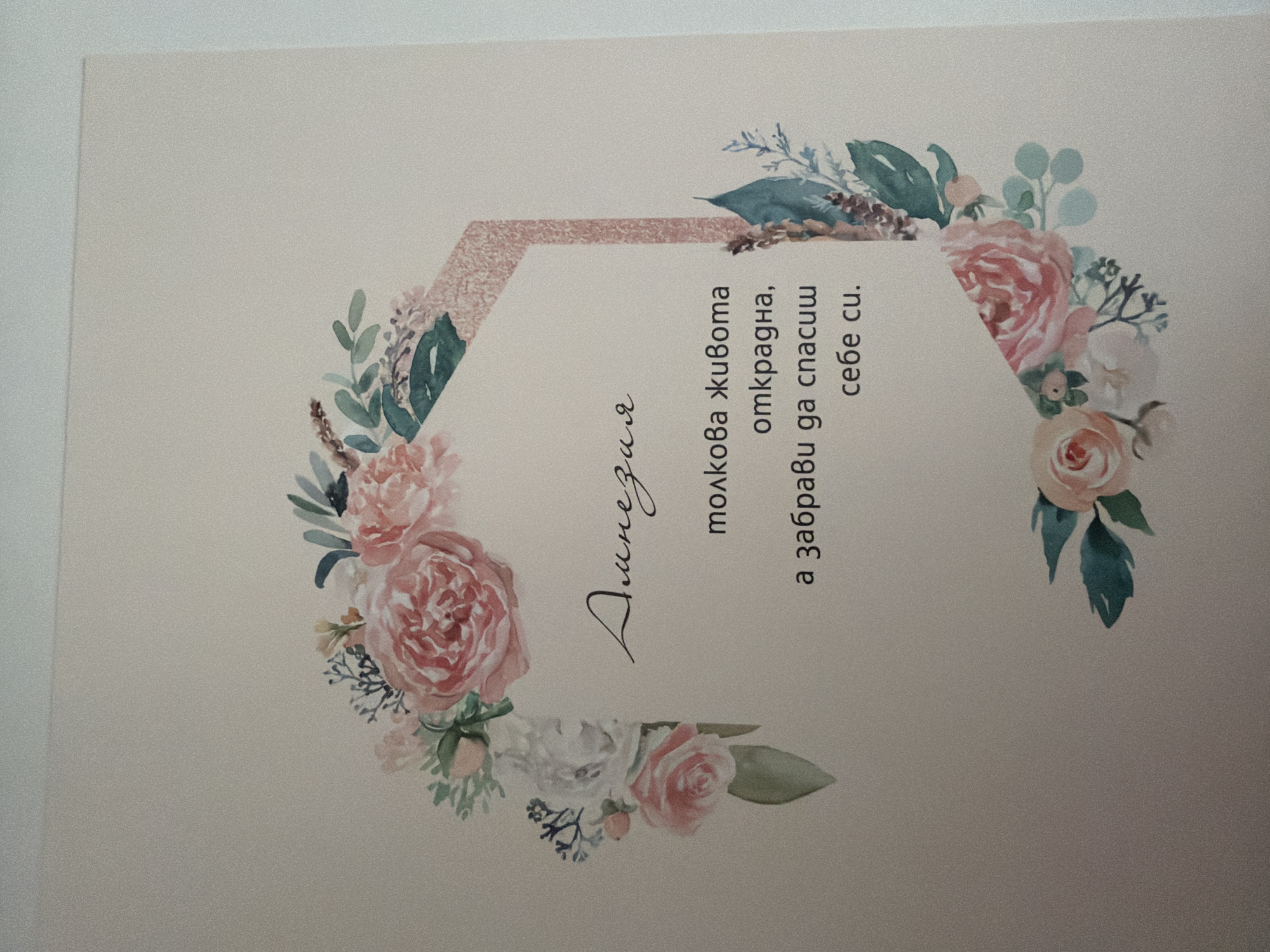 The image is a close-up, eye-level shot of a greeting card with a floral design. A wreath of watercolor-style pink and white roses, green leaves, and small blue and purple accents encircles a hexagonal frame. Inside the frame, in black font, is text in Bulgarian. The top line, in a script font, reads "Динездия". Below this, in a serif font, are three lines of text: "толкова живота", "открадна,", and "а забрави да спасиш себе си.". The background of the card is a soft, pale beige. The lighting is even, and the focus is sharp on the card. No external environment or location information is present in the image.