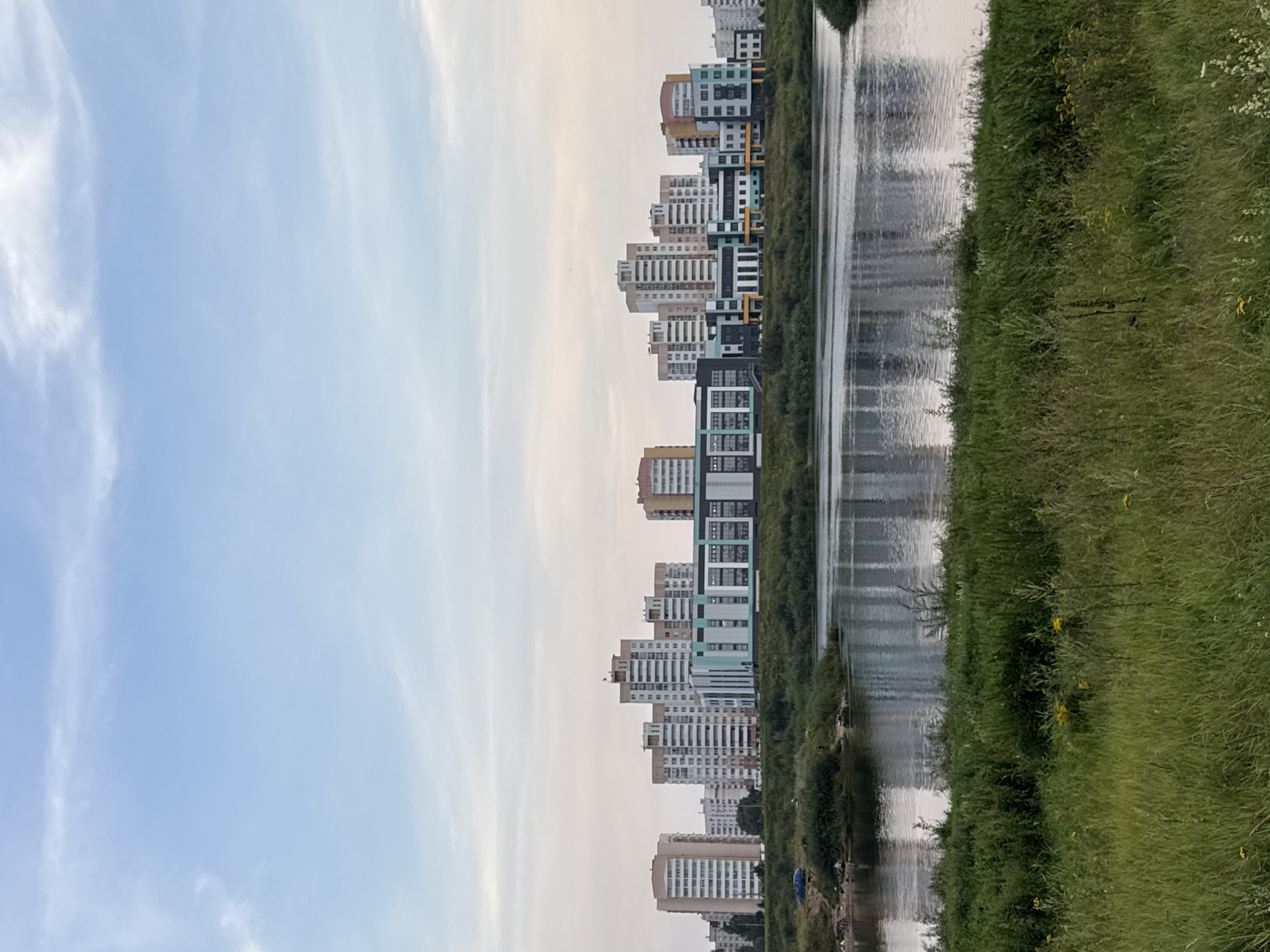 An outdoor vertical shot depicts a body of water in the foreground with a city skyline in the background. The water is calm and reflects the buildings and sky. The foreground is dominated by lush green grass. In the middle ground, a riverbank is covered in dense green vegetation. On the far bank of the river, a line of modern, multi-story buildings stands against a pale, hazy sky with thin clouds. The buildings appear to be apartment complexes and possibly a school, featuring a mix of light and darker colored facades. Some of the buildings have geometric architectural details, and one building in the center has a particularly vibrant turquoise and green color scheme. To the left of the buildings, a small sandy area on the riverbank has a few people and a blue car. The overall impression is a contemporary urban development situated alongside a natural water feature. This scene is likely located in Mahilyow, Belarus, a city known for its proximity to the Dnieper River and its modern architectural projects.