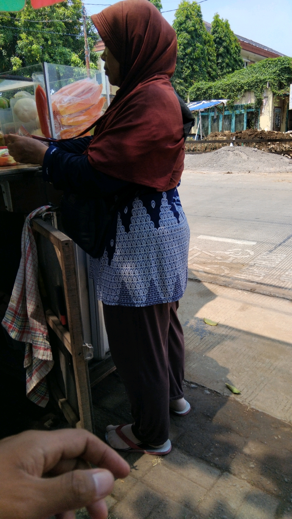 In Batang, Indonesia, a woman wearing a brown hijab and a dark blue top with white patterned detailing stands next to a food stall. She is holding something in her hands, likely serving a customer. The stall displays sliced fruits, including papaya, in a clear container. A checkered cloth hangs from a wooden support near the stall. The woman is wearing dark pants and flip-flops. The background shows a street with a sidewalk, some vegetation, and buildings, one of which appears to be under construction or renovation. The lighting suggests it is daytime. A person's hand is visible in the foreground, partially obscuring the lower part of the image.