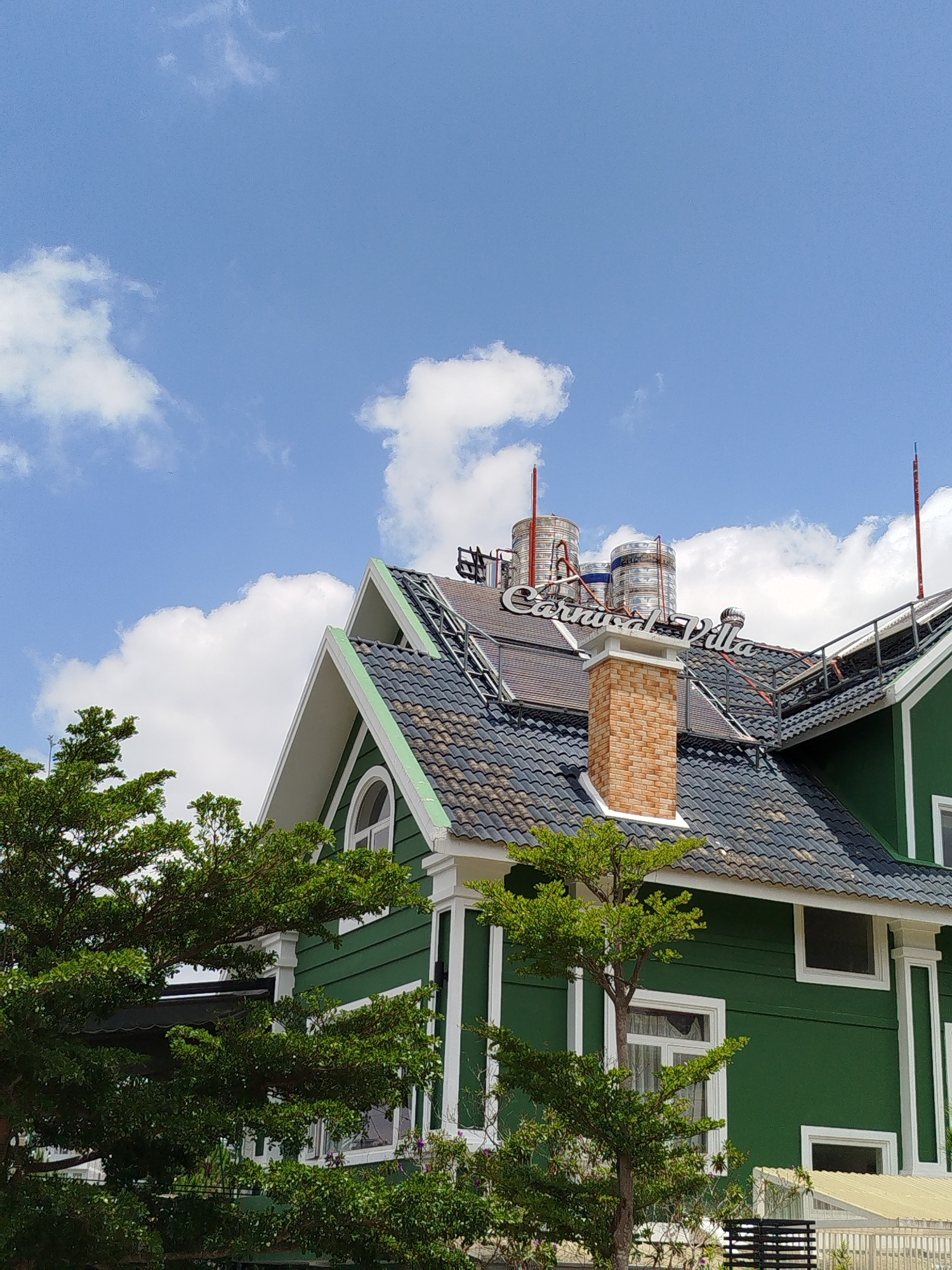 An outdoor, eye-level shot captures a vibrant green villa or house under a bright blue sky with scattered white clouds. The building features a distinctive green facade with white trim outlining its windows, gables, and architectural details. Its dark-tiled roof is equipped with several prominent features, including large, cylindrical stainless steel water tanks, what appear to be solar panels, and a red-brick chimney. White, raised lettering on the rooftop clearly spells "Carnival Villa." Tall, lush green trees partially obscure the lower and left portions of the building, providing natural framing. The sky is a clear, brilliant blue, indicating bright sunshine and fair weather, likely during the mid-morning or early afternoon. No people or specific activities are visible, emphasizing the building and its natural surroundings. The scene depicts a picturesque property, characteristic of locations like Da Lat, Vietnam.