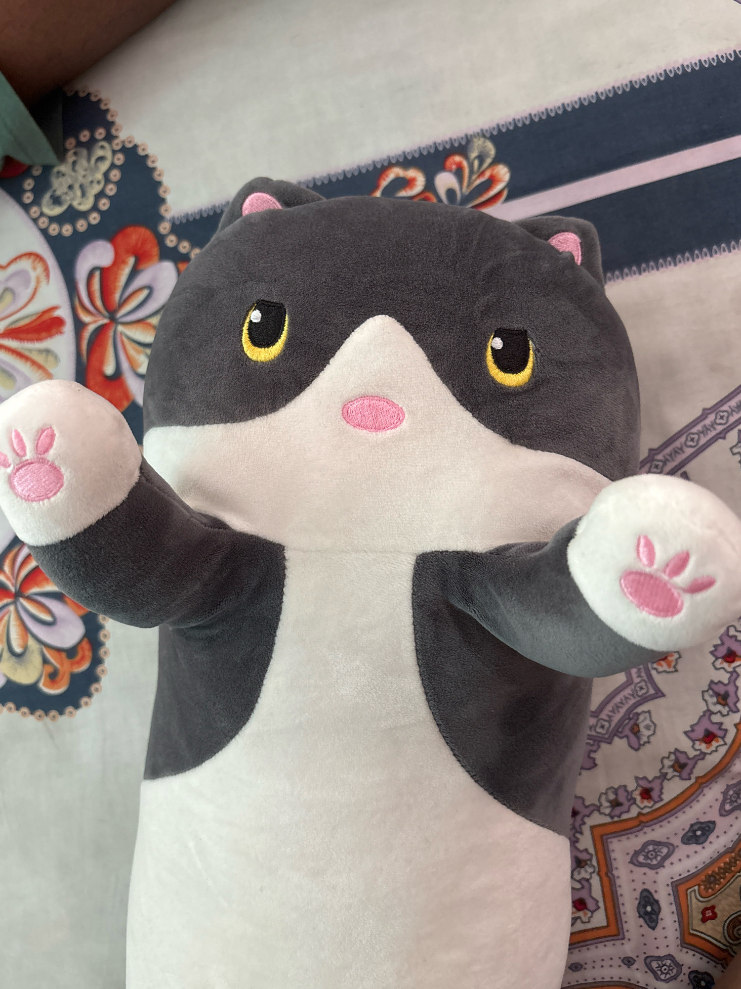 A plush toy cat is lying on a bed with a patterned sheet. The cat is mostly gray with white on its chest and paws. It has yellow eyes, pink ears, and a pink nose. The sheet has a blue and white floral pattern. The image is taken from a top-down perspective. The location of this image is not discernible from the provided visual information.