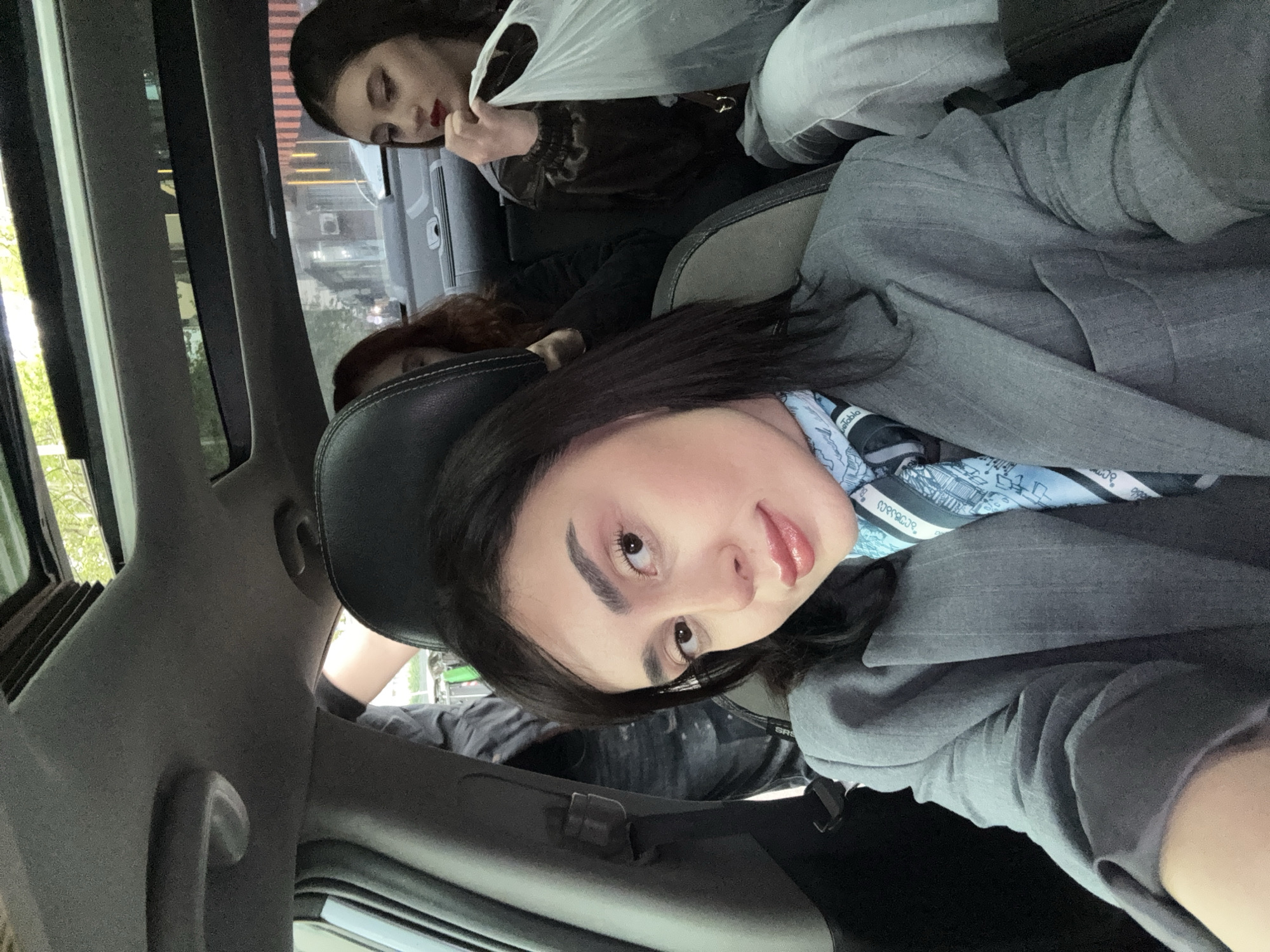 The image depicts three women inside a car. One woman in the foreground is wearing a grey blazer and a blue scarf. Another woman behind her is holding a white plastic bag. A third woman with red hair is behind the first. The location is Tbilisi, Georgia, based on a storefront sign visible through the window. The car's interior is grey.