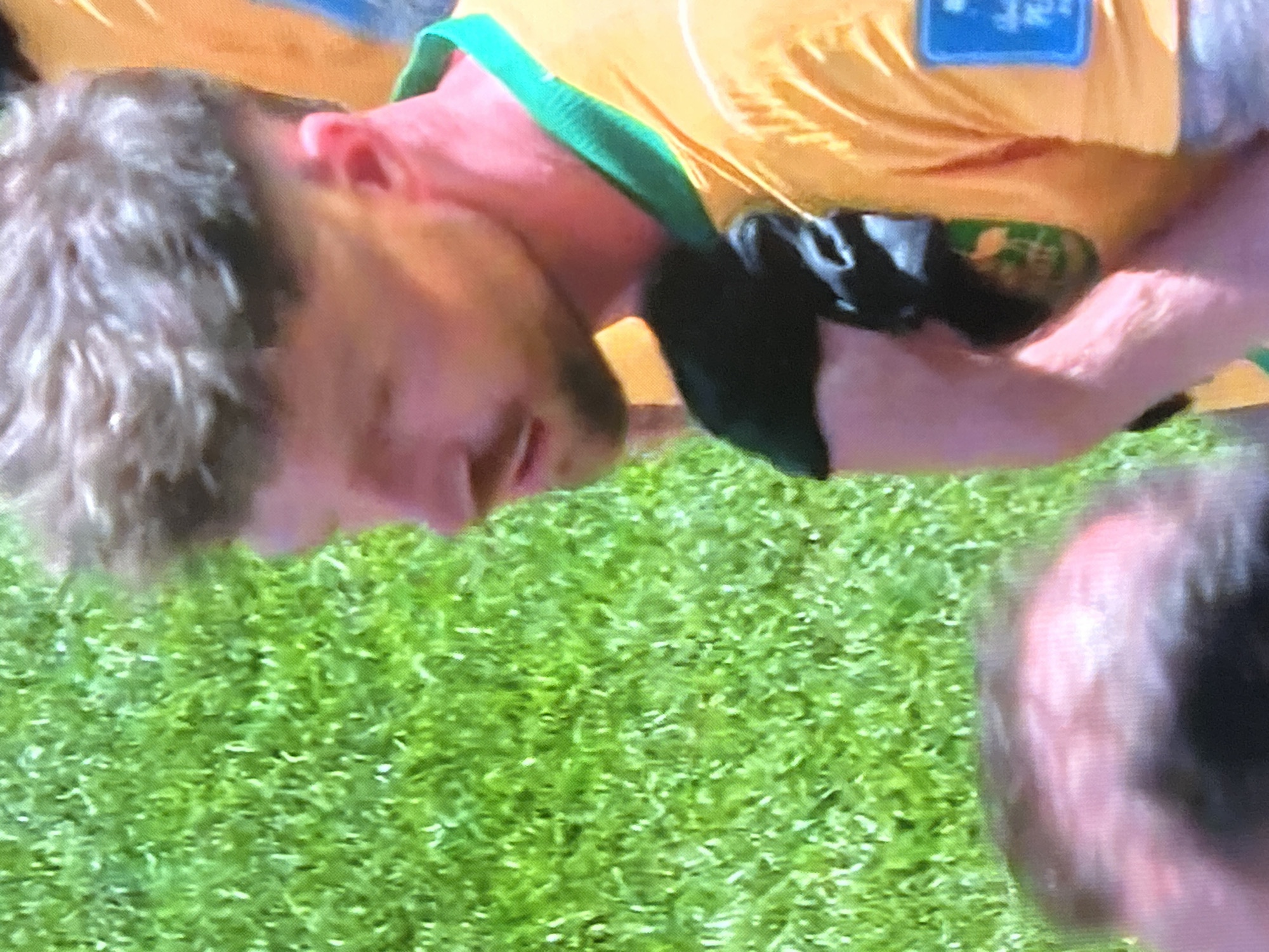 The image is a close-up, slightly angled shot of a light-skinned man with short, spiky brown hair, a short beard, and a slight stubble. He is wearing a bright yellow sports jersey with a green collar. His skin appears flushed, and there are visible sweat beads on his neck and upper chest. He is looking downwards and to the left, with his mouth slightly open. He is holding a dark-colored piece of fabric in his left hand, which is partially obscuring his chest. In the background, a blurred green surface, likely grass, is visible. Another person's head, with dark hair, is partially visible in the lower left corner of the frame. A blue rectangular patch with white text is affixed to the right shoulder of the man's jersey. The lighting suggests an outdoor environment. The image quality is somewhat grainy. The location is Unknown, Unknown.