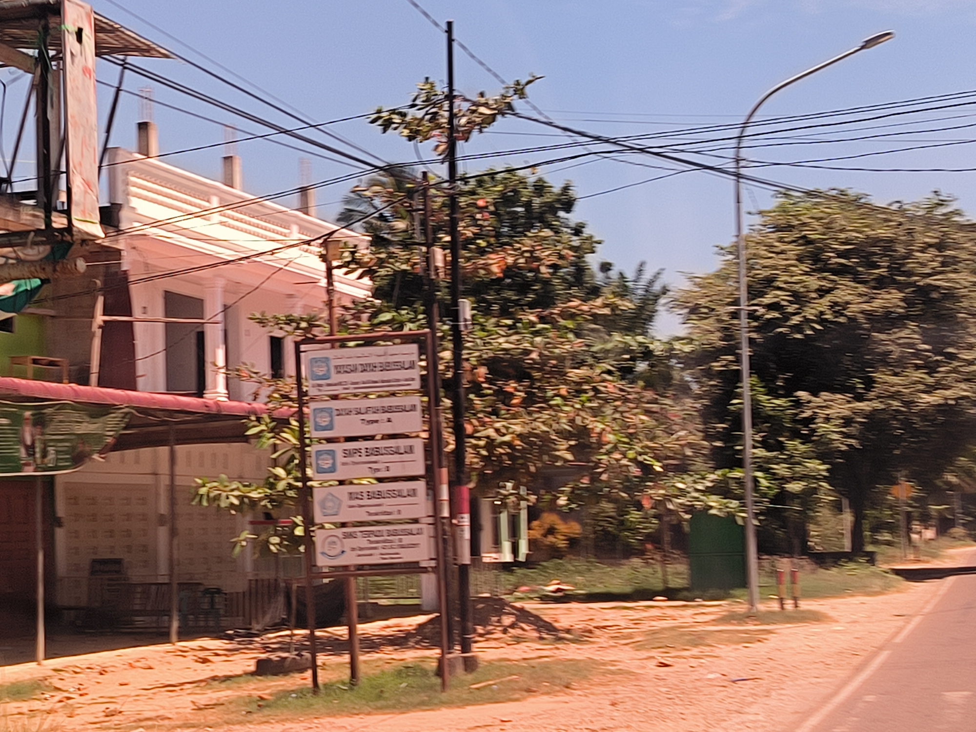 The image shows a street scene in Keudealue Ie Puteh, Indonesia. On the left is a building with a partly-covered porch or patio area. Adjacent to it is a signpost displaying multiple signs, possibly indicating the location of educational or institutional facilities. Power lines are strung overhead. A street light stands to the right of the signpost. Trees and foliage are visible in the background. The road appears to be unpaved in some areas. The sky is clear and blue. No people are discernible in the frame.