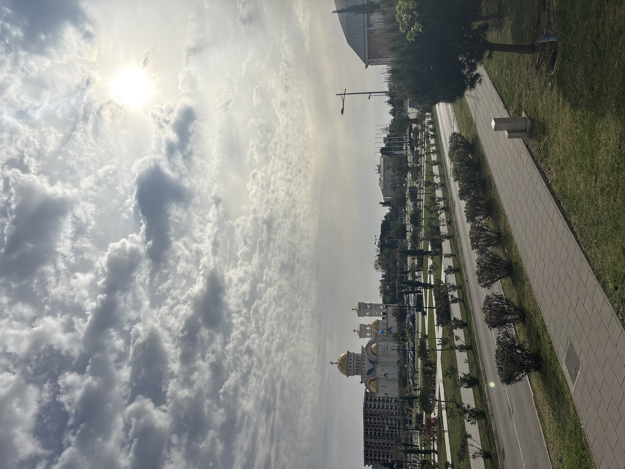 The image captures a cityscape in Bar, Montenegro, under a partly cloudy sky with the sun visible. The prominent feature is the Temple of Saint Jovan Vladimir, an Orthodox church with golden domes, located adjacent to a modern apartment building. A long, straight road lined with trees and streetlights extends into the distance. A gray tiled walkway runs parallel to the road, bordered by grass. A white utility box is situated near the sidewalk. In the background, other buildings and urban structures are visible.