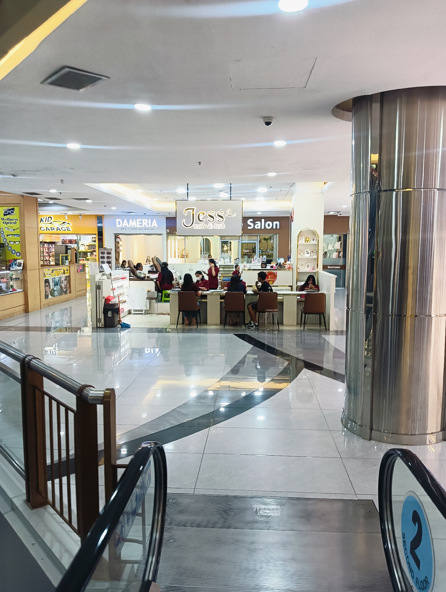 The image shows the interior of a shopping mall in Batam, Indonesia. In the foreground, a silver, cylindrical pillar is prominently visible on the right. An escalator with a black handrail and a blue sign indicating "SECOND FLOOR" is on the lower right. The floor is made of polished white tiles with dark grey diagonal lines. In the midground, there are several shopfronts. A nail and lash salon named "Jess2 Nails & Lash" is in the center. To its left is a shop called "DAMERIA" and to the right is a shop called "SALON". Several people are seated at counters in front of the salons, appearing to be receiving services or waiting. Some employees are wearing red uniforms. Further to the left, a shop labeled "KID GARAGE" is visible, displaying merchandise. The ceiling has recessed lighting and ventilation grates.