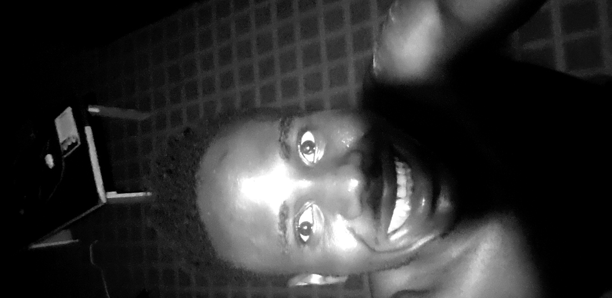 A close-up, low-resolution, monochrome image features a person's face, slightly out of focus, with a visible smile and eyes directed toward the camera. The person appears to have a bald head. The background consists of a patterned surface, possibly a tiled wall or floor. An object resembling a small table or shelf with some items on it is visible in the left portion of the frame. No evidence indicates it is in Narayi, Nigeria.