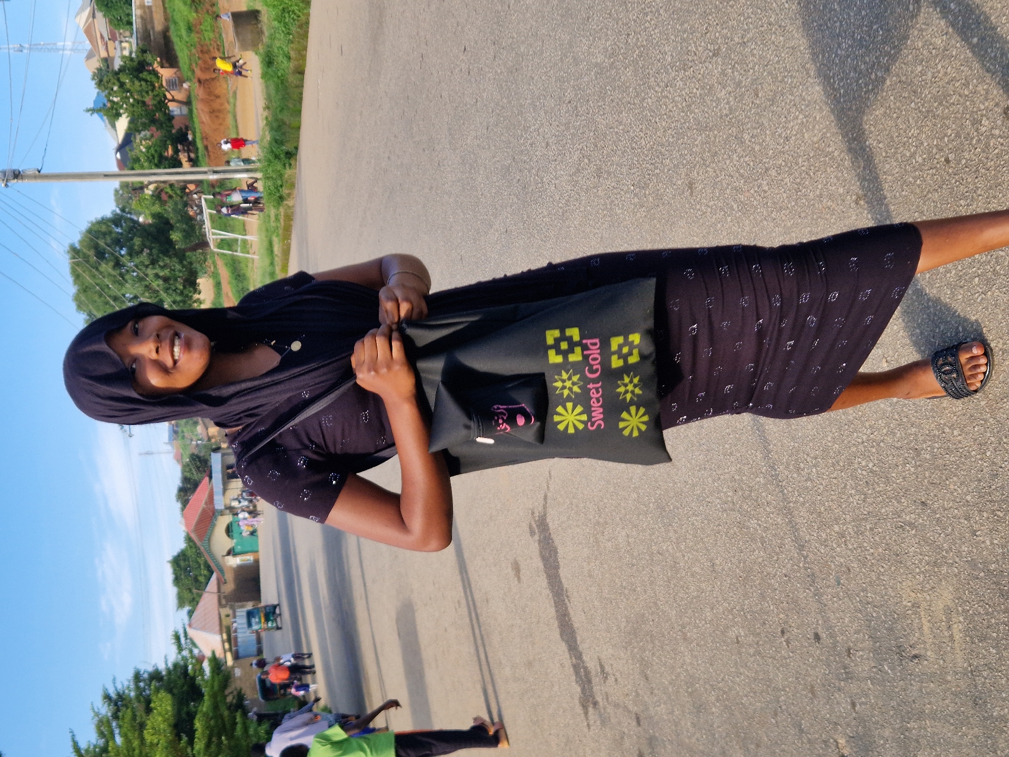 A Black woman wearing a black hijab and a patterned black dress walks down a paved road in Kabong, Nigeria. She carries a black tote bag with yellow floral designs and the words "Sweet Gold" printed on it. In the background, several people are walking and interacting, and a small green auto-rickshaw is visible. Buildings with red and green roofs are present, along with trees and utility poles. The sky is clear and blue, with some clouds. A soccer goal is visible on the right side of the frame.
