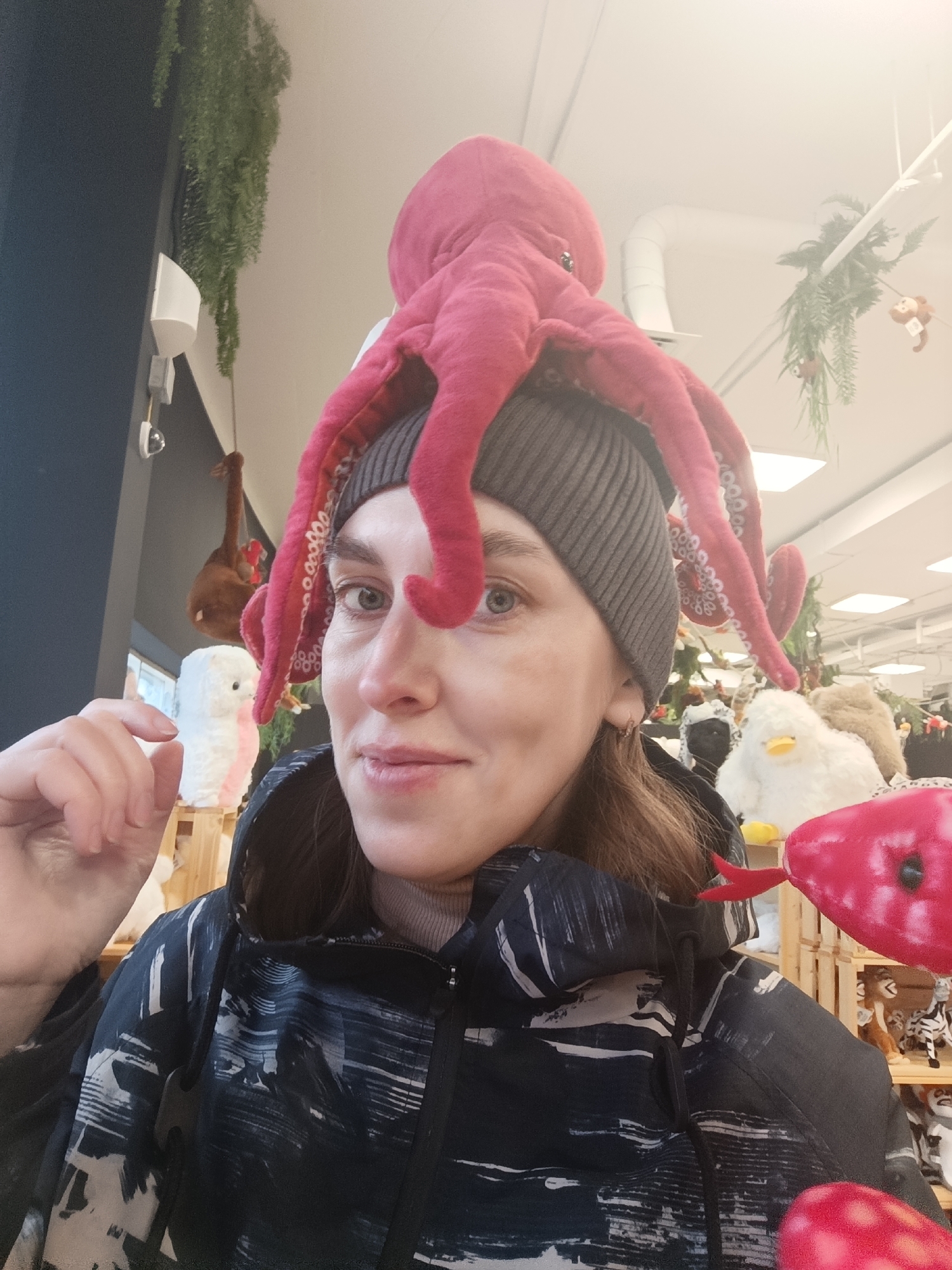A fair-skinned woman with light eyes and brown hair is wearing a gray knit hat topped with a red stuffed octopus. She is also wearing a black and white patterned jacket. The photo appears to have been taken indoors, possibly in a store, as shelves of stuffed animals are visible in the background. Decorations, including hanging plants, are also visible in the background. The photo may have been taken in the city of Perm, Russia.