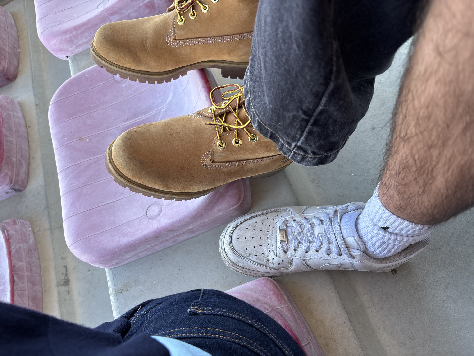 The image shows a ground-level shot of two people sitting in a stadium. The person on the left is wearing dark blue jeans and Timberland boots. The person on the right is wearing black jeans and a white Nike Air Force 1 shoe with a white sock. They are sitting on light purple stadium seats. The location is presumed to be in the city of Valpaços, Portugal.