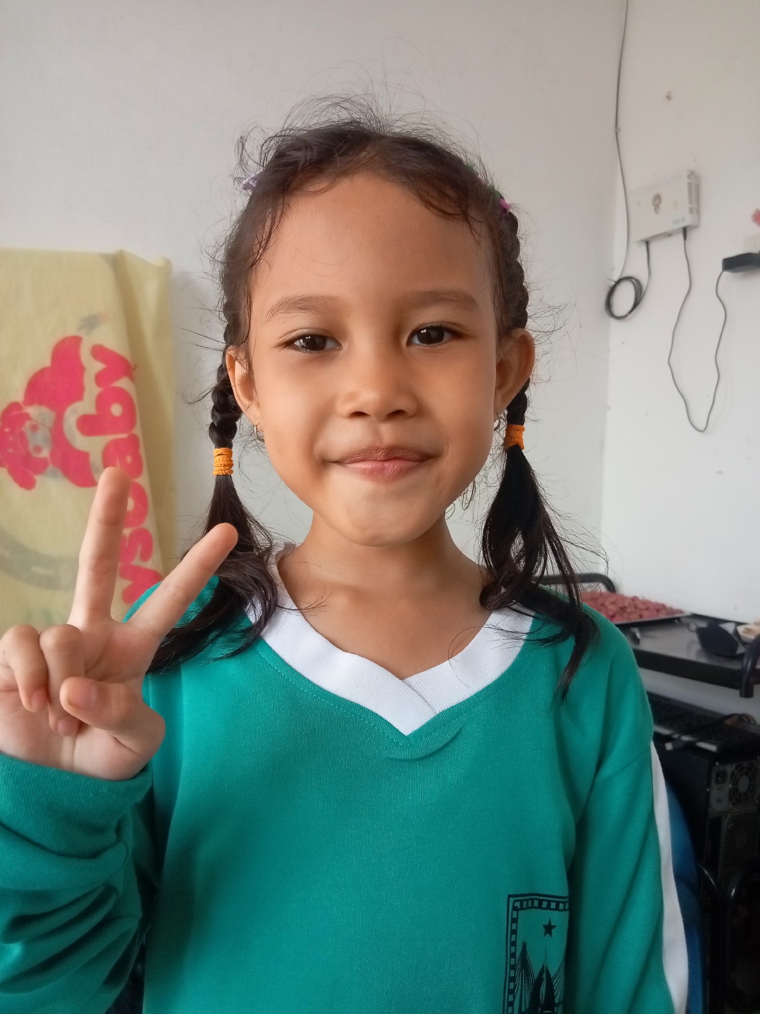 A young girl with dark hair in two braids, tied with orange bands, and wearing a teal long-sleeved shirt with a white collar, smiles at the camera and holds up a peace sign with her left hand. She is standing indoors against a white wall. To her left, a yellow cloth with a pink graphic is visible. To her right, electrical outlets and wires are mounted on the wall, and a dark object, possibly a computer tower, is partially visible in the background. The setting appears to be a residential area in Batam, Indonesia.