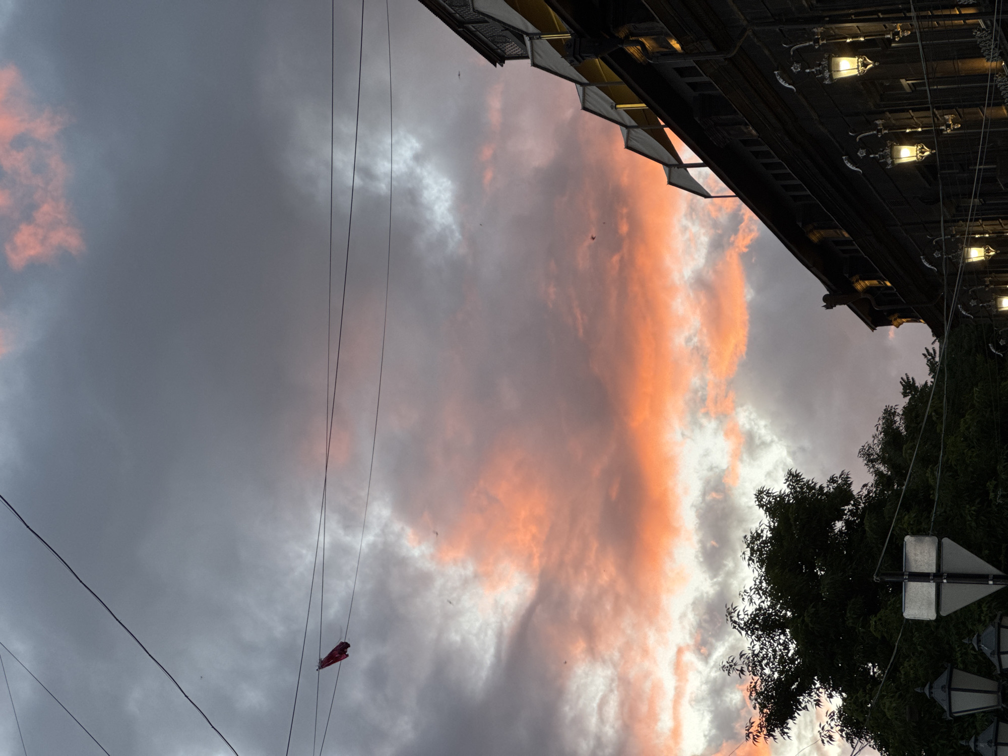 The image captures an outdoor scene in Yerevan, Armenia, featuring a dramatic sky with pink and grey clouds, framed by urban elements. Power lines crisscross the sky. At the top right, there's a fragment of a building with awnings and illuminated outdoor lights. The lower right corner contains the leaves of a tree and a traffic sign. In the lower left portion of the sky, a small red kite hangs tangled in the power lines. The photo appears to be taken from street level.