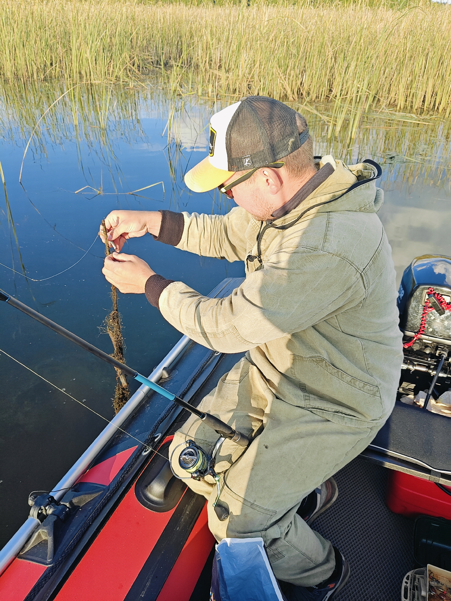 A man wearing a tan hooded jacket, a trucker hat with a yellow brim, and sunglasses is sitting in a boat. He is holding a fishing lure in his hands, examining it. A fishing rod is resting in the boat next to him. The boat is on a body of water with tall reeds lining the shore. A small outboard motor is attached to the back of the boat. The scene appears to be in Chaykovsky, Russia, based on the context.