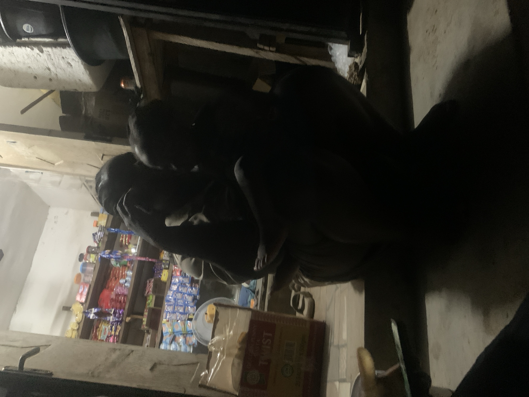 The image depicts a dimly lit scene in Lokoja, Nigeria, seemingly inside or at the entrance of a small shop. Shelves filled with various snacks and grocery items are visible in the background. A partially visible electric fan and a carton labeled "TWIST" sit in the foreground. Two individuals are huddled close to each other. The poor lighting obscures their features, but their posture suggests an intimate interaction or private conversation.