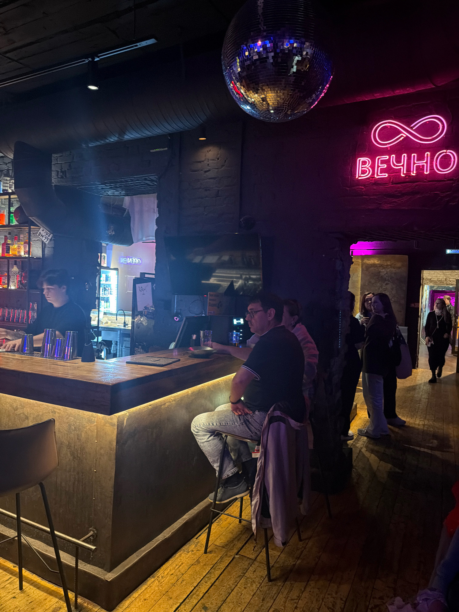 The image depicts the interior of a dimly lit bar in Samara, Russia. A bartender, wearing a black t-shirt, is visible behind a wooden counter, appearing to be preparing drinks. Several patrons are present, including a man seated at the bar wearing glasses, a black t-shirt, and jeans, with a jacket draped over his chair. Another group of individuals stands further back in the bar area. A prominent feature of the bar's decor is a neon sign displaying an infinity symbol and the word "ВЕЧНО" (eternally) in pink, mounted on a brick wall. A disco ball hangs from the ceiling. The environment is characterized by exposed brick, wooden flooring, and ambient lighting, contributing to a nightlife atmosphere.
