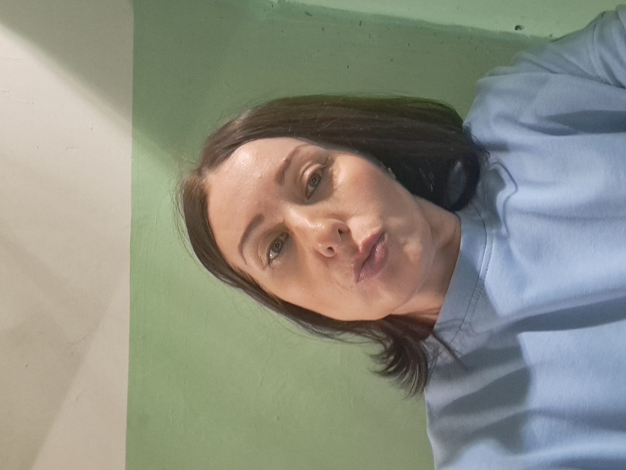 A woman with dark brown hair is pictured from the chest up, looking at the camera with her lips puckered. She is wearing a light blue sweatshirt. The background is a wall painted in two shades of green, with a lighter shade at the top and a darker shade below. The lighting suggests an indoor environment. There are no identifiable landmarks or features that would indicate a specific location within the city of Irkutsk, Russia.