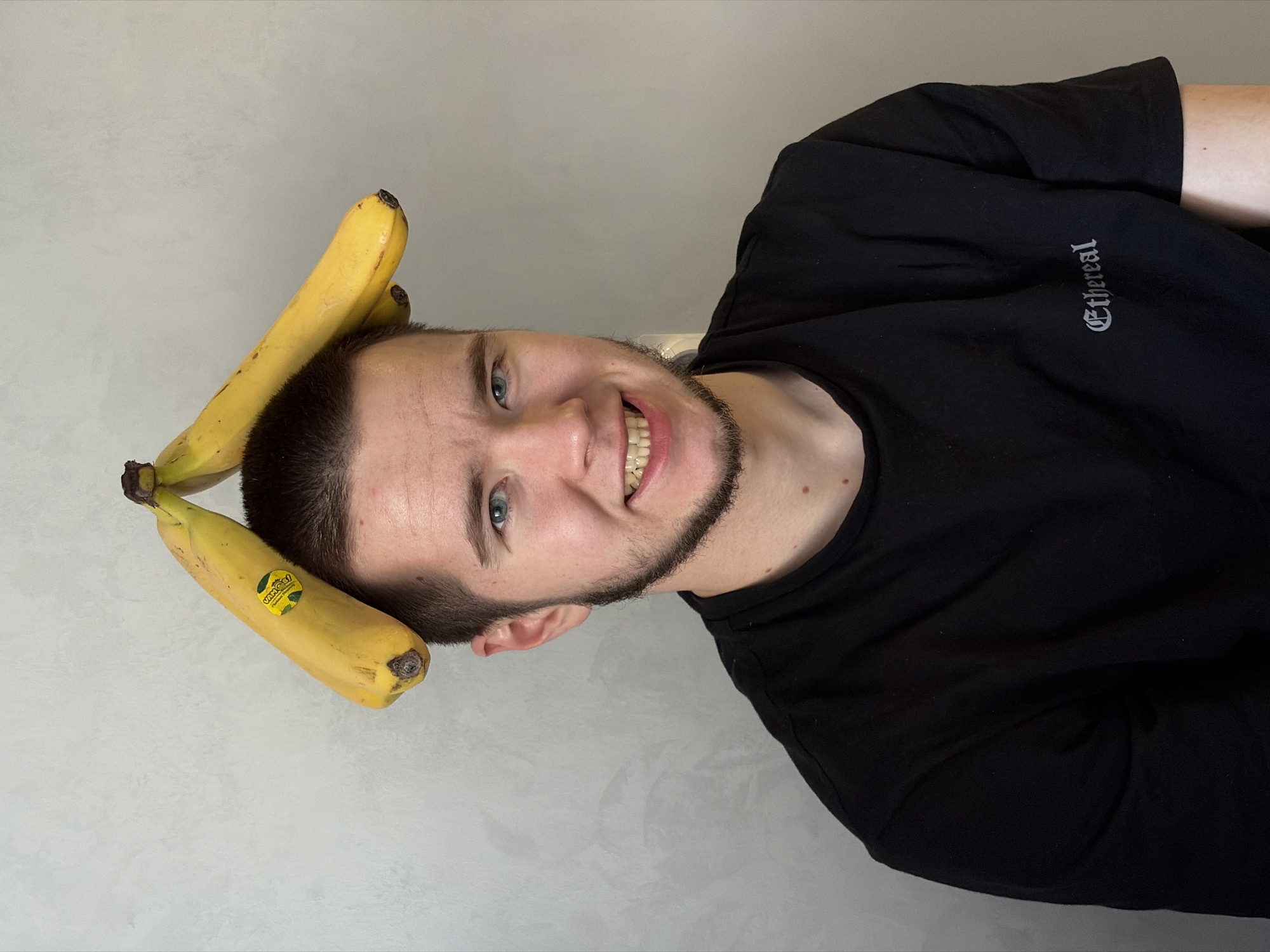 A Caucasian male with short dark hair and a beard is smiling at the camera. Two yellow bananas are placed on his head, forming a triangle shape. He is wearing a black t-shirt with the word "Ethereal" printed in a gothic-style font. The background is a plain, light grey wall. The image shows no indication it was taken in Sarapul, Russia.